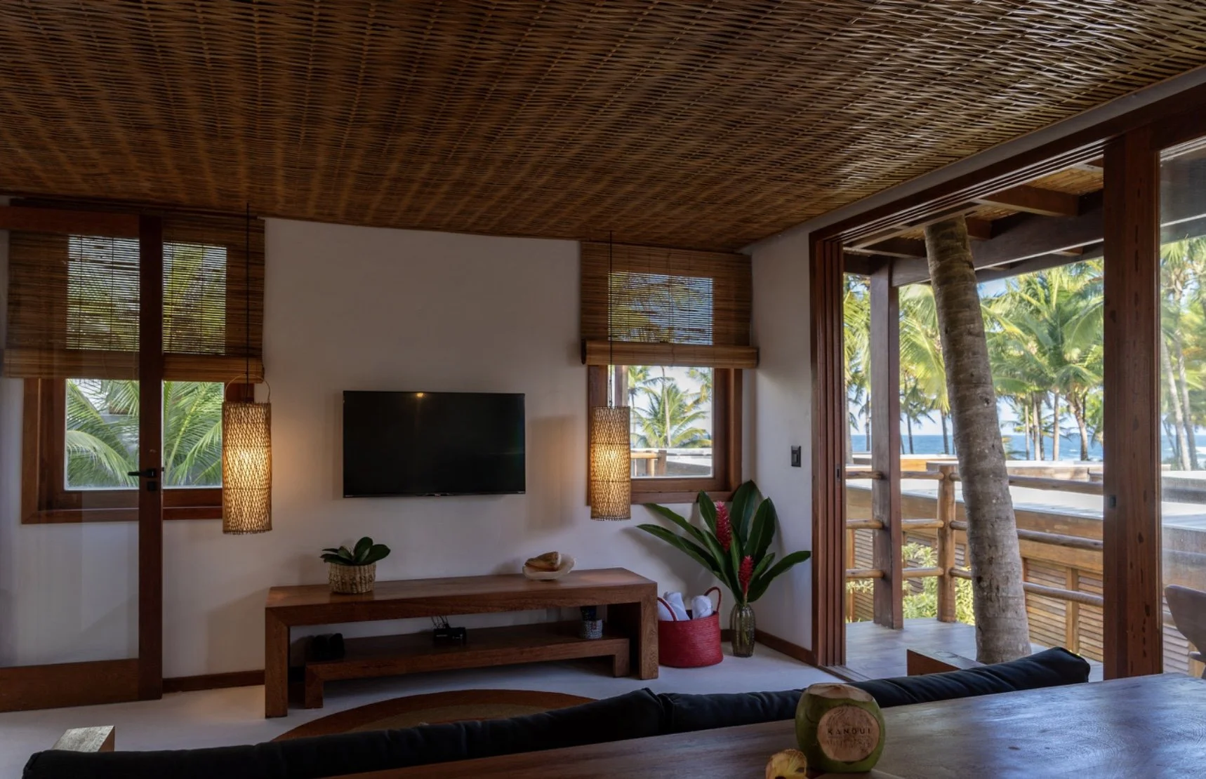 Sala de estar decorada com estilo rústico, com janela e porta de vidro que abre para uma varanda com vista para árvores tropicais e o mar, decorada com plantas, luminárias de fibra natural, TV e mobília de madeira.