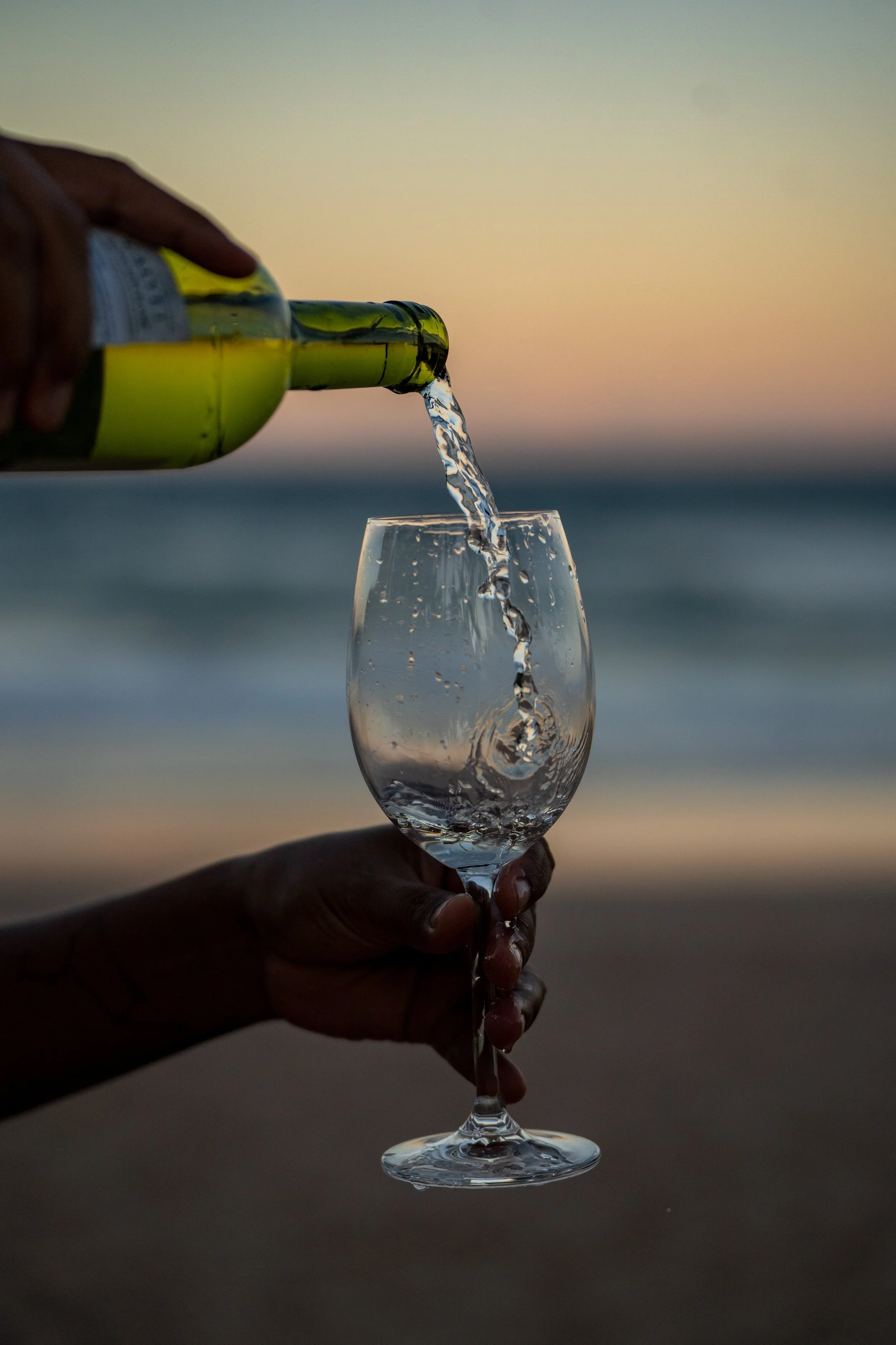 Pessoa serve água de garrafa de vidro em uma taça na praia ao pôr do sol.