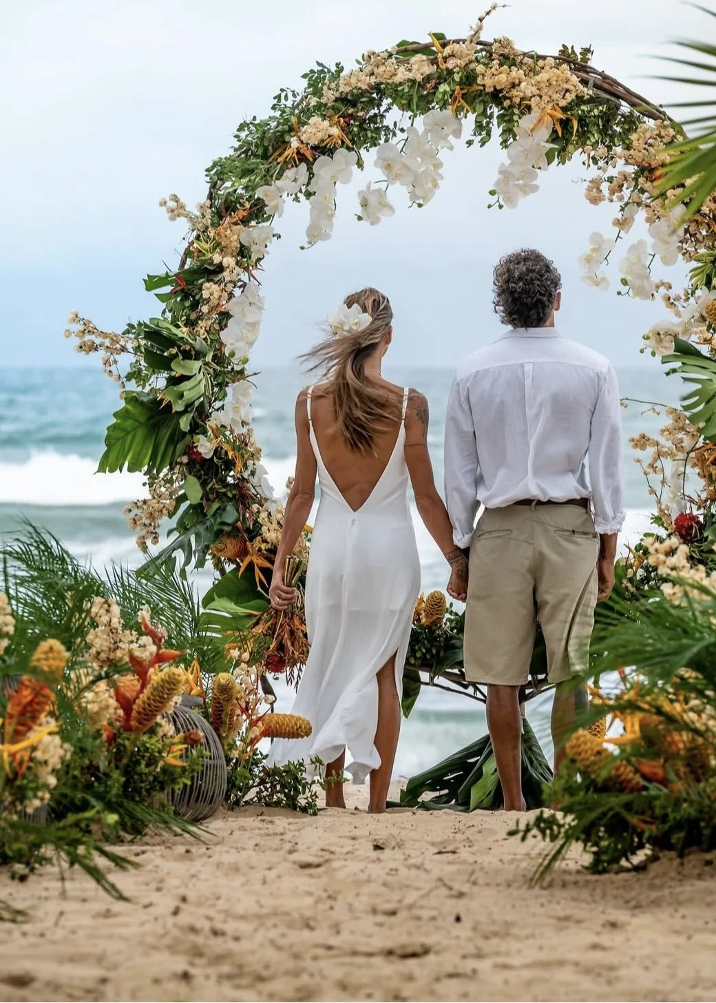 Casal de casamento na praia sob arco decorado com flores e plantas tropicais, vista para o mar ao fundo