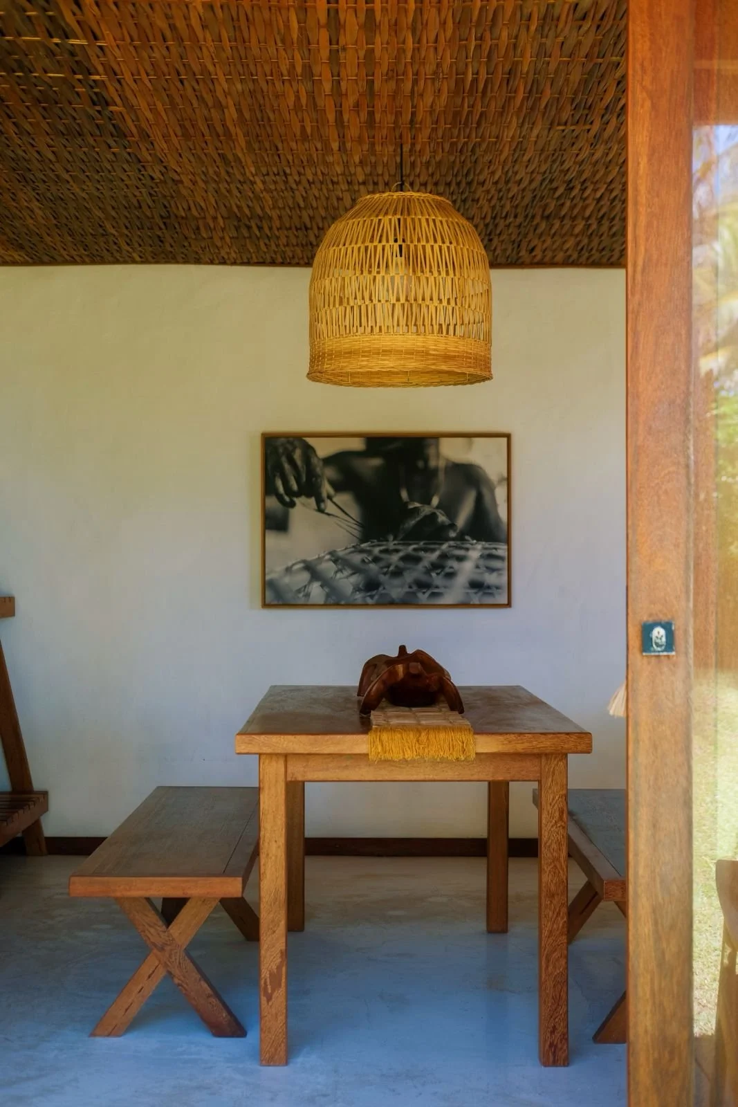 Sala de jantar com mesa de madeira simples, bancos de madeira, luminária de fibra natural suspensa, quadro preto e branco na parede e uma escultura de madeira sobre a mesa.