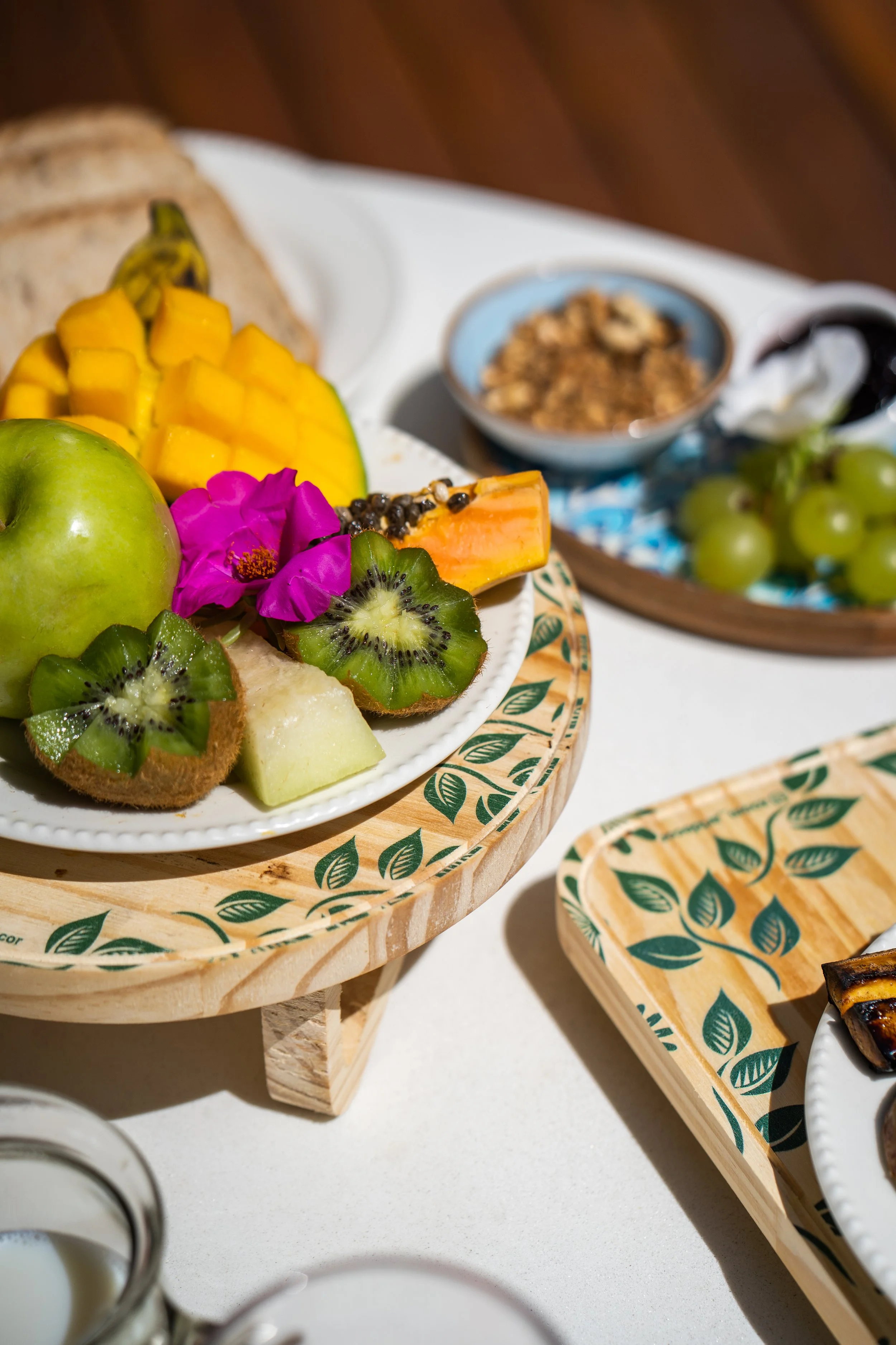 Banquete de café da manhã com frutas variadas, incluindo kiwi, maçã verde, manga, mamão e uvas, decorado com uma flor rosa. Há também pão e granola ao fundo, servidos em pratos e bandejas com design de folhas.