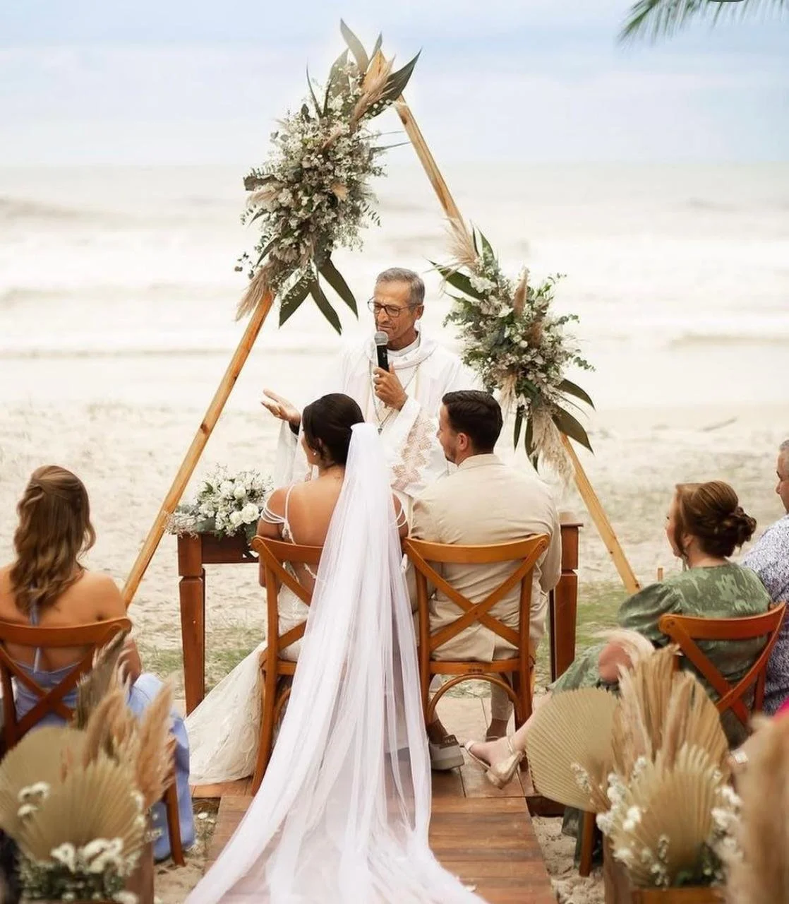 Casamento na praia com altar decorado com flores e folhagens. No altar, um casal e um oficiante. Os convidados assistem ao casamento ao redor.