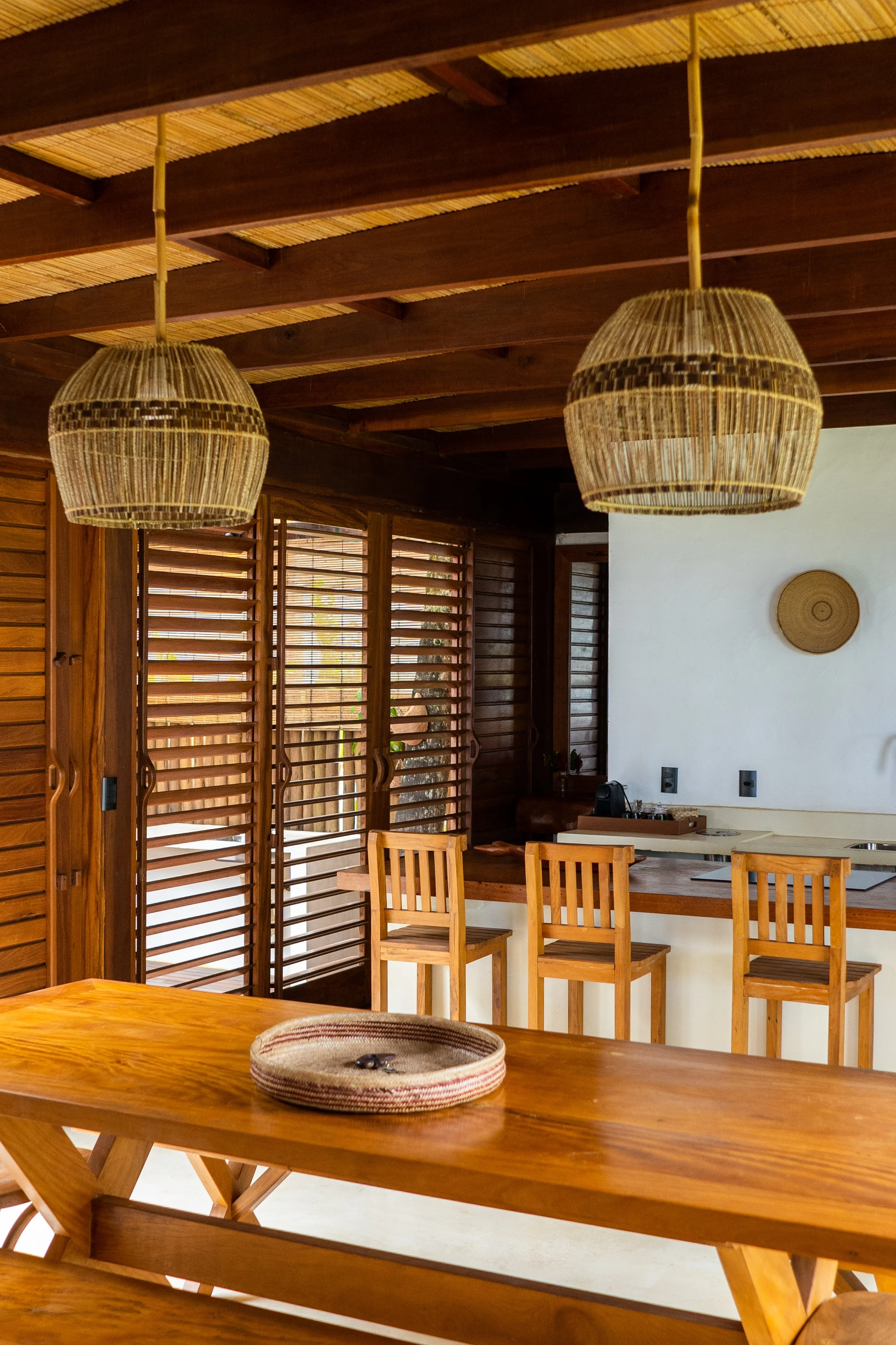 Interior de uma cozinha com móveis de madeira, duas luminárias de fibra natural suspensas, persianas de madeira nas janelas e uma bancada com três cadeiras de madeira.