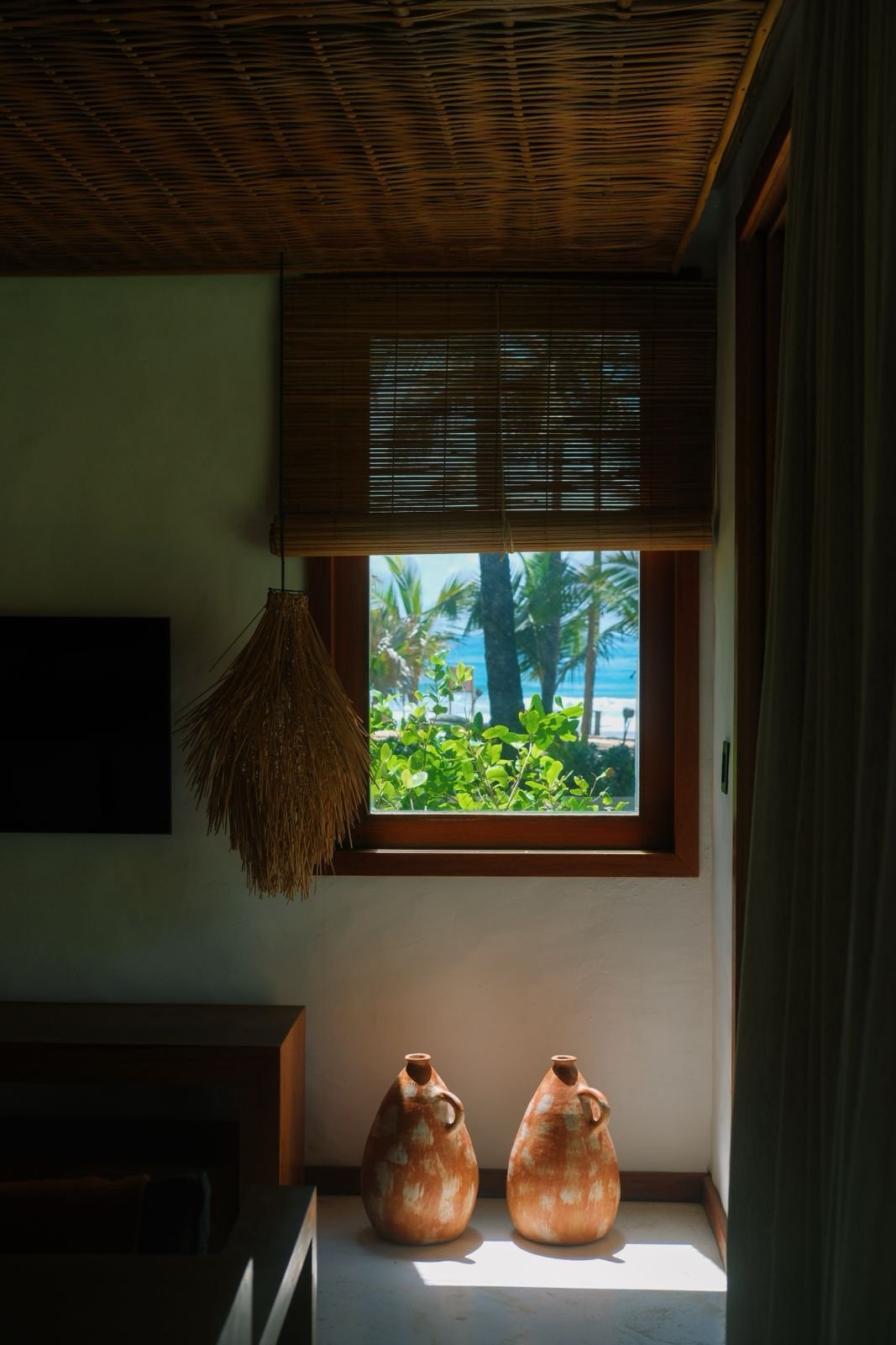 Janela de madeira com vista para palmeiras e praia, cortinas de bambu, luminária de fibra natural e duas urnas de cerâmica decorativas no piso