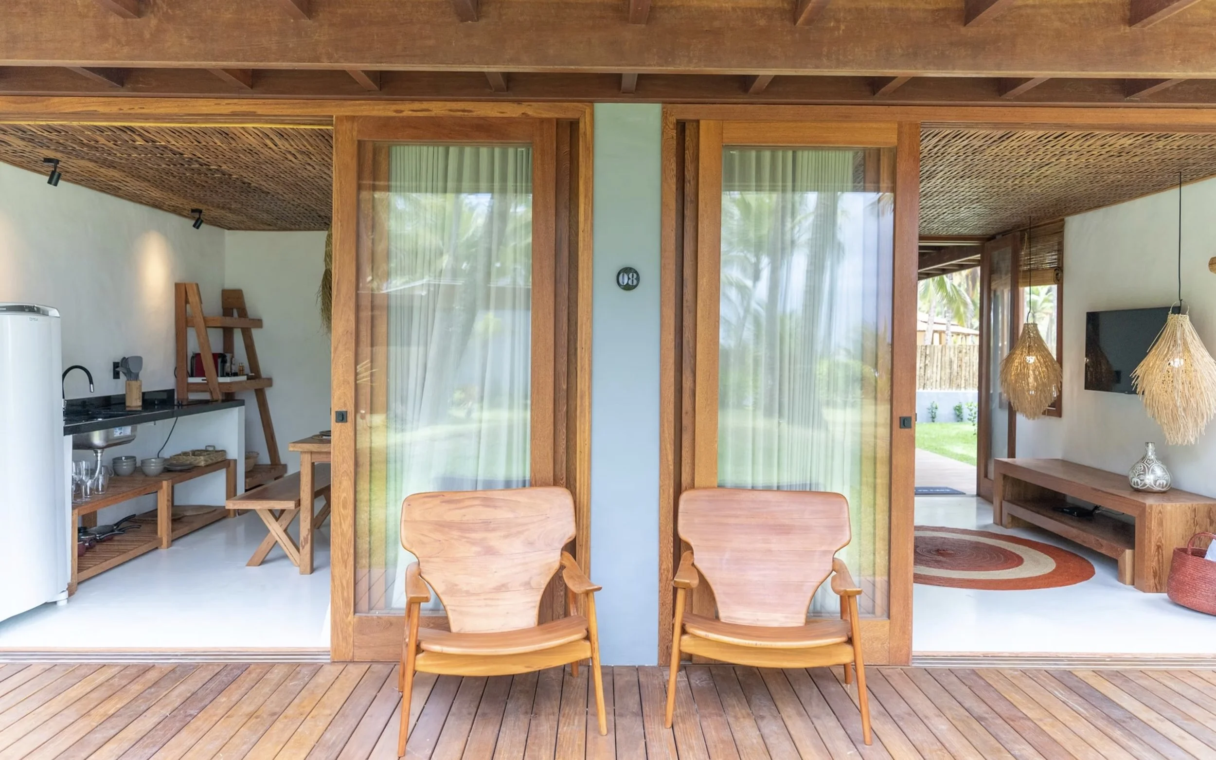 Varanda de casa de madeira com duas cadeiras de madeira no primeiro plano, sala com móveis de madeira, televisão, luminárias de fibra natural, tapete redondo e vidro com cortina branca na casa.