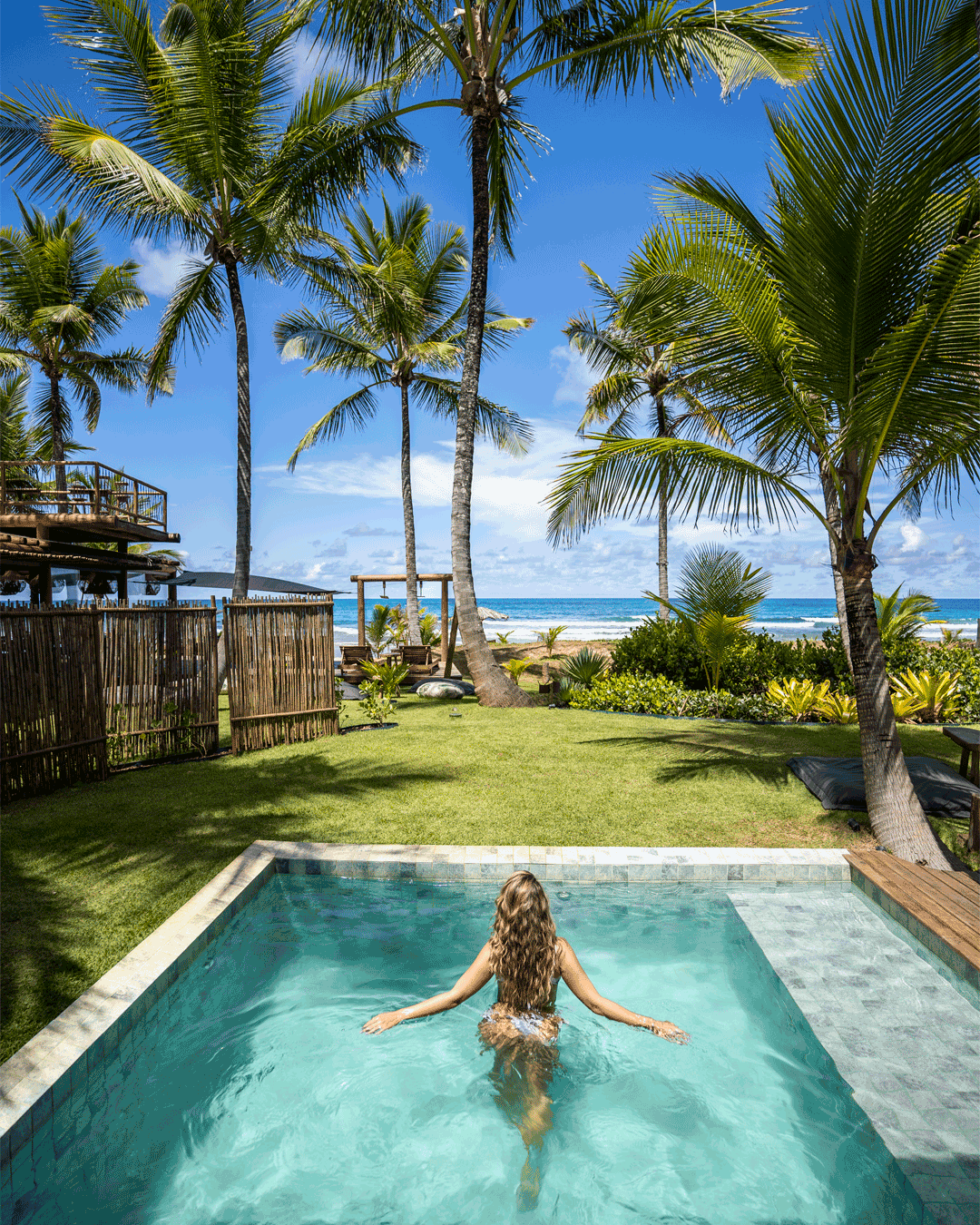 Pessoa com cabelo cacheado na piscina, cercada por árvores de palmeira, com praia e oceano ao fundo.