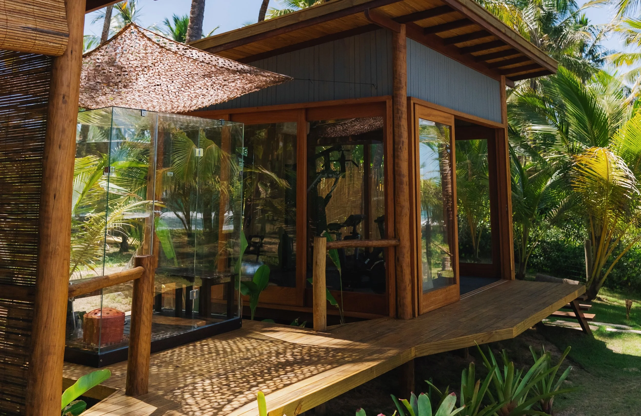 Uma estrutura de cabana de madeira com paredes de vidro, localizada em um ambiente tropical com muitas plantas verdes e Palmeiras ao redor, sob um céu azul claro.