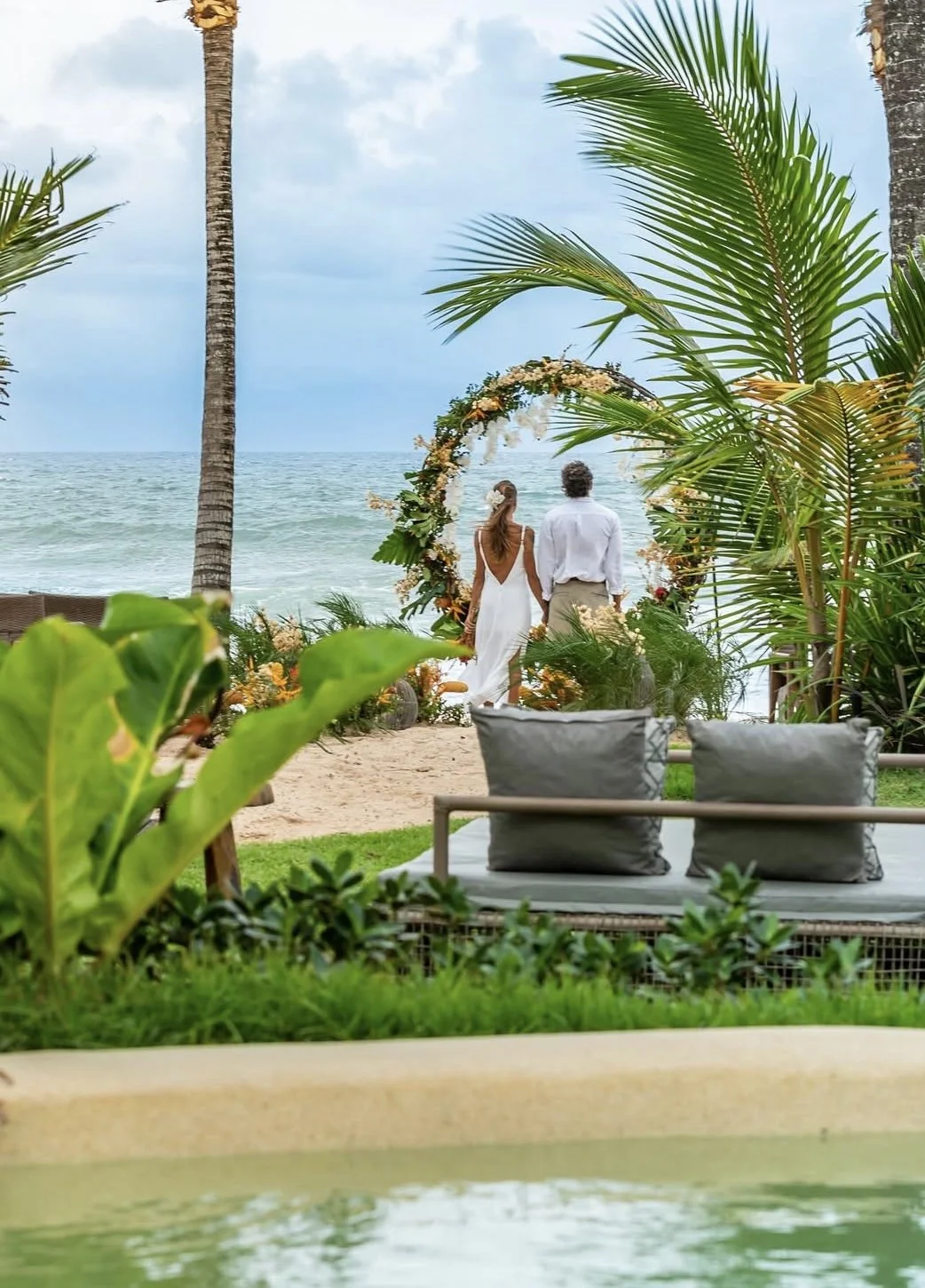 Casal de noivos assistindo ao pôr do sol na praia, com decoração de arco de flores, rodeado por plantas tropicais, em ambiente de casamento ao ar livre.