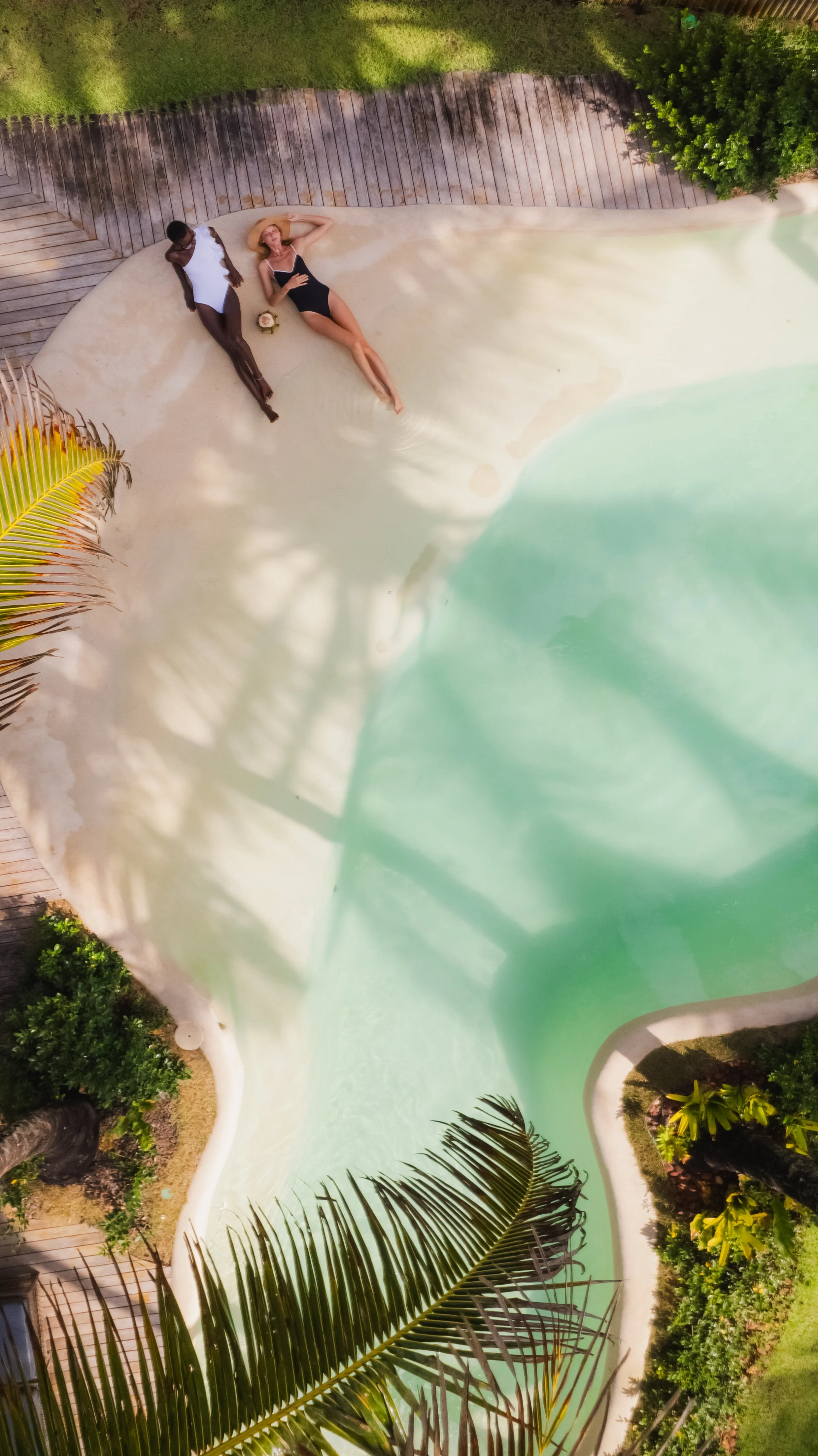 Cena aérea de duas pessoas descansando à beira de uma piscina, cercada por vegetação tropical.