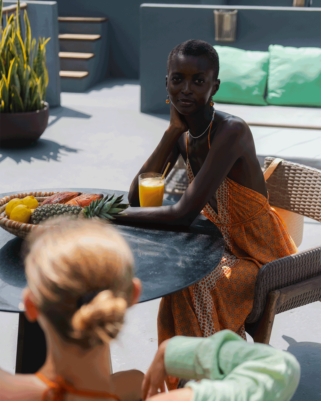 Mulher com pele escura e cabelo curto, usando vestido laranja com estampa, sentado à mesa com uma bebida de suco de laranja, na varanda ao ar livre. A mesa tem frutas como abacaxi, limões e manga, com uma mulher de cabelos claros e penteado preso na 