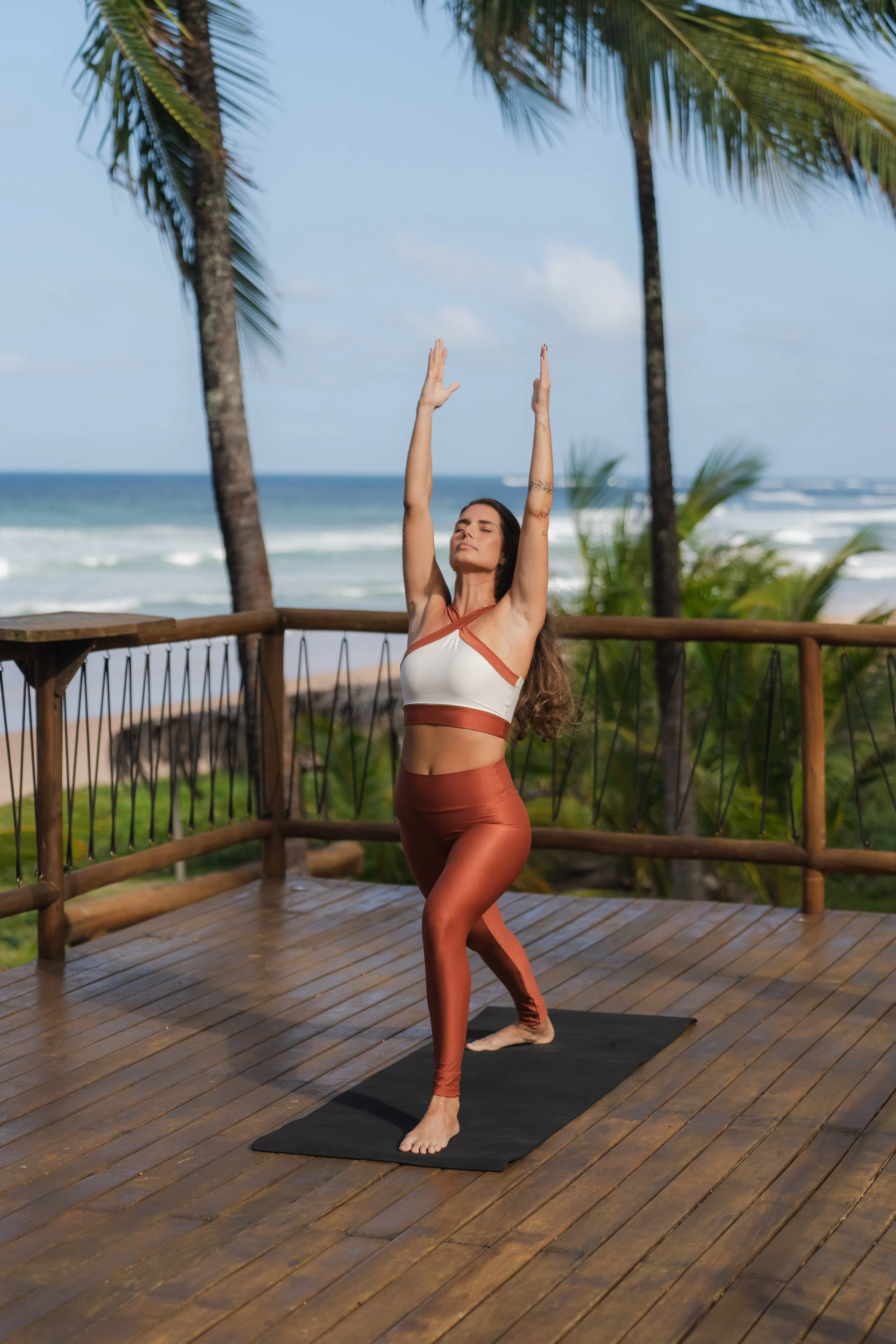 Mulher fazendo yoga em terraço de madeira com vista para o mar e coqueiros, usando roupa de ginástica marrom e branca.