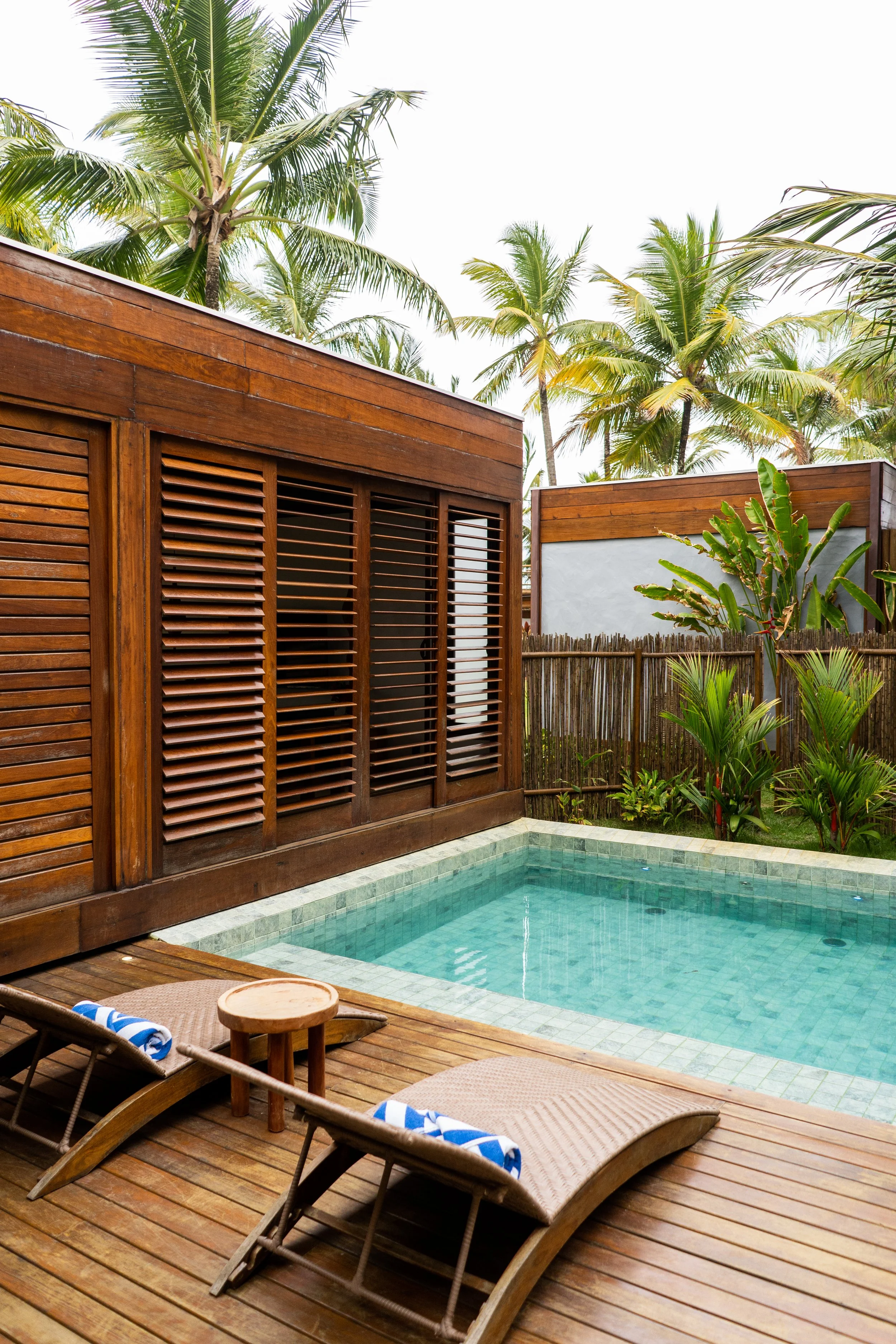 Área externa com piscina, espreguiçadeiras de madeira com toalhas azuis e brancas, palmeiras ao fundo, e parede de madeira com venezianas.
