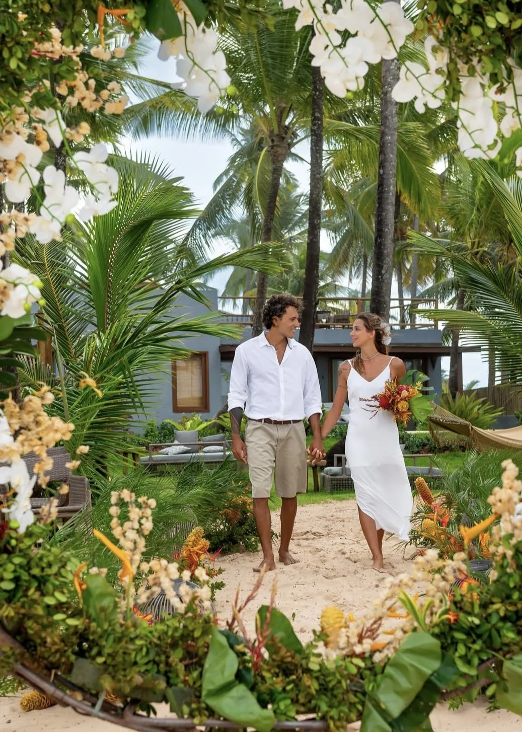 Casal de noivos caminhando de mãos dadas em um cenário de casamento ao ar livre, cercados por plantas tropicais e flores, com uma casa moderna ao fundo.