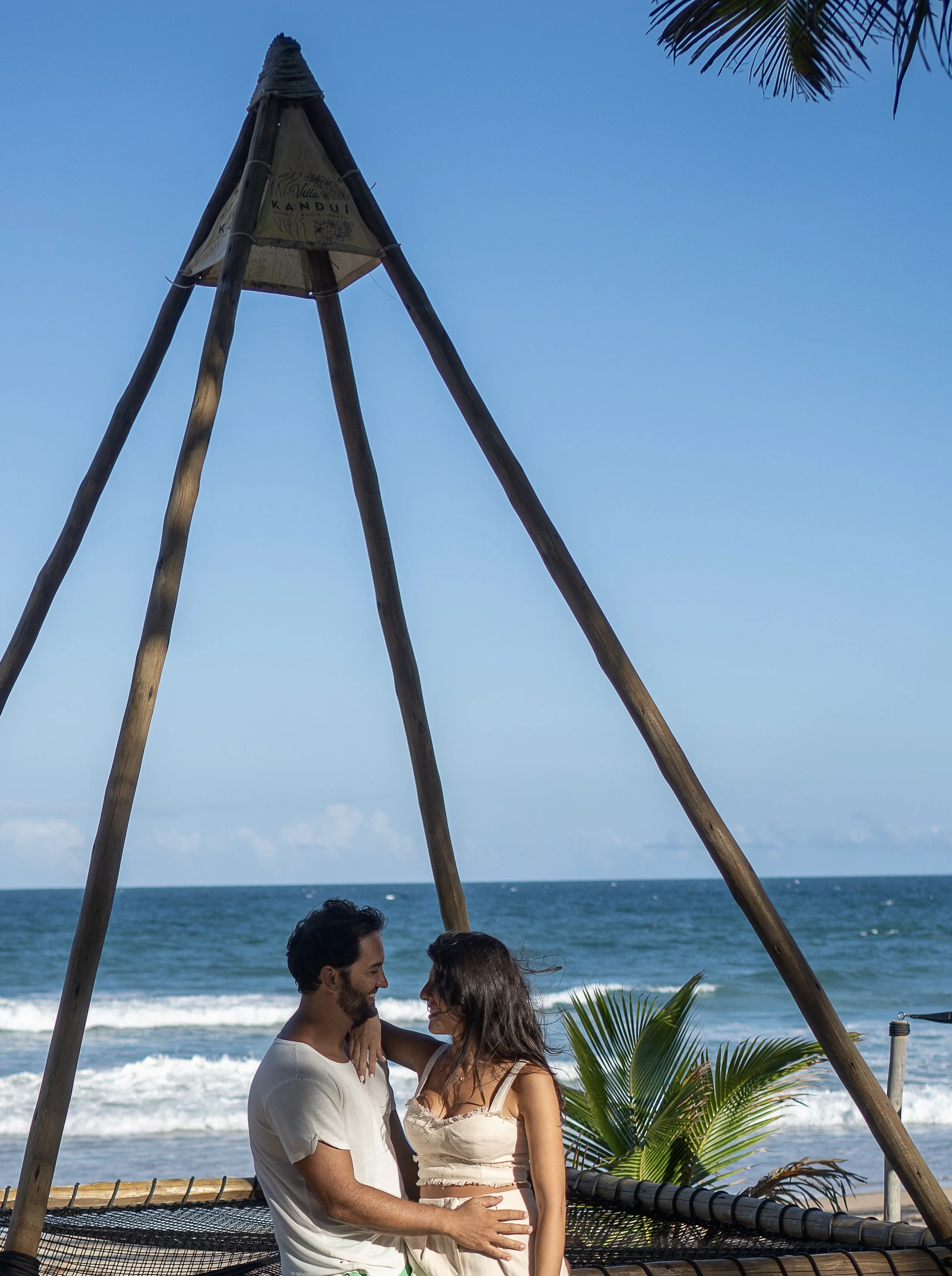 Casal sorridente na praia, com vista para o oceano, sob uma estrutura de madeira com cobertura de palha e plantas ao redor.