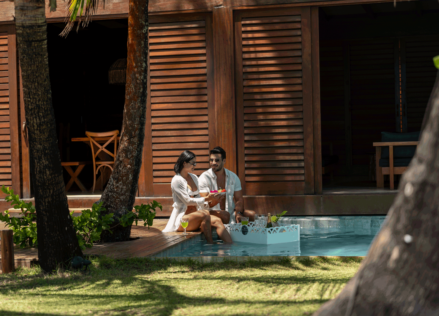 Casal relaxando junto à piscina em um ambiente ao ar livre, com uma estrutura de madeira ao fundo e árvores ao redor, durante o dia.