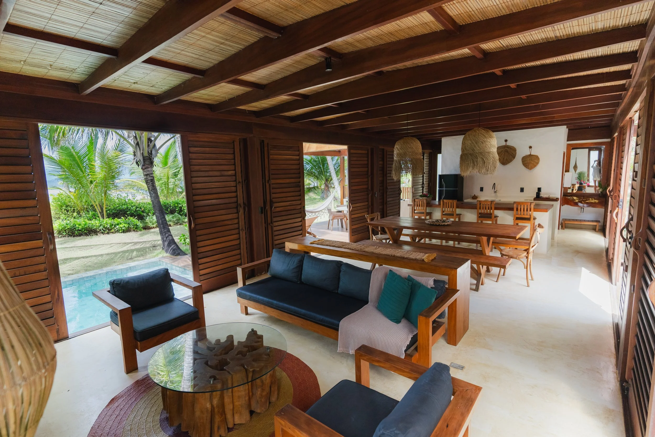 Sala de estar de uma casa de estilo rústico com móveis de madeira, sofá preto, cadeiras, mesa de centro de madeira, parede de madeira, vista para jardim com piscina e vegetação tropical.