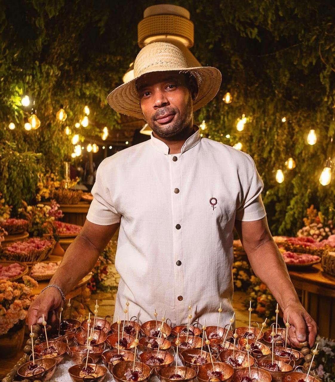 Homem com chapéu de palha, veste clara, em ambiente decorado com luzes e flores, segurando pratos com doces.