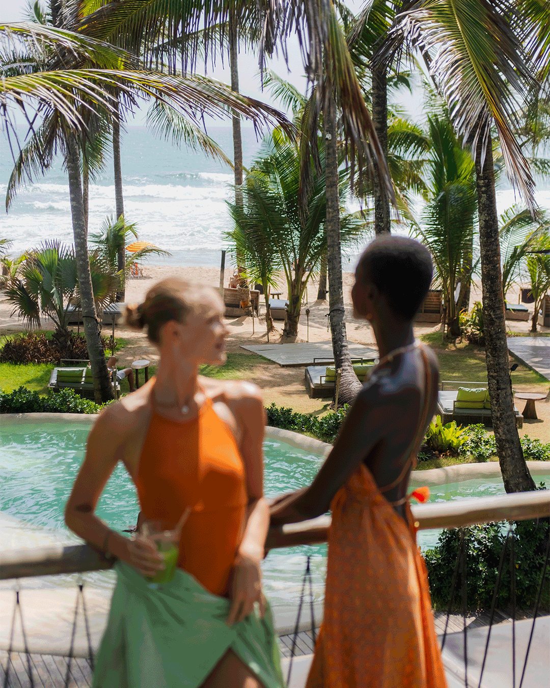 Duas mulheres conversando perto de uma piscina em uma área com palmeiras na praia, com o mar ao fundo.
