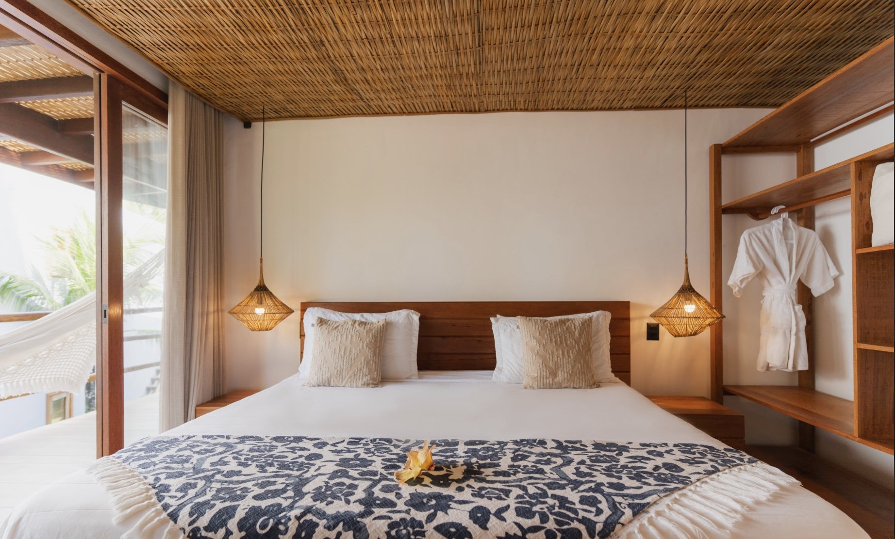 Quarto de hotel com cama de casal, decoração de estilo rústico, paredes brancas, cabides de luz pendurados, varanda com poltrona de descanso, vista de plantas ao lado.