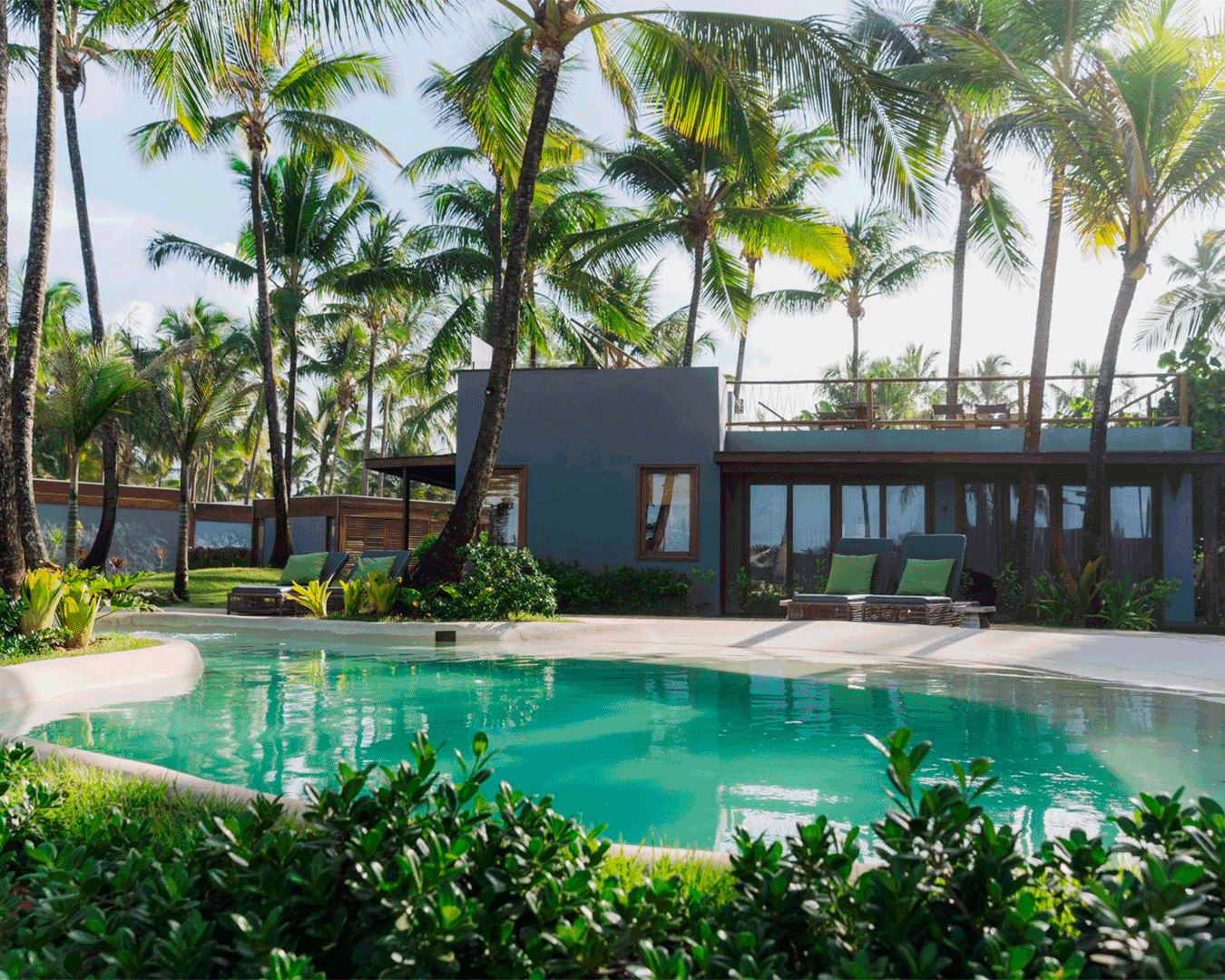 Piscina ao ar livre rodeada por vegetação tropical e palmeiras, com uma casa moderna ao fundo. Móveis de relaxamento estão próximos à piscina.