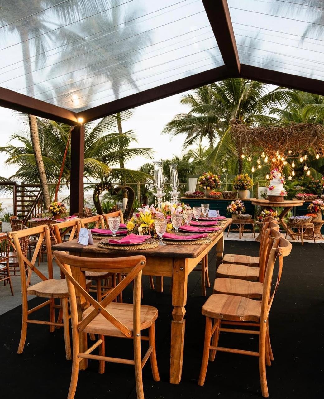 Mesa decorada com flores, taças e pratos em um ambiente ao ar livre com palmeiras ao fundo, preparado para uma festa ou celebração.