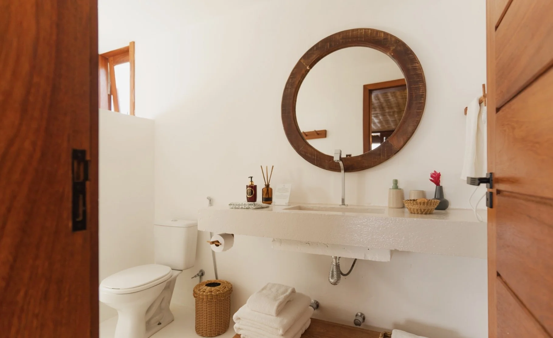 Banheiro com espelho de moldura de madeira, pia de pedra, vaso sanitário e decoração com itens como sabonete líquido, difusor, vasilha com flores e toalhas dobradas em uma prateleira.