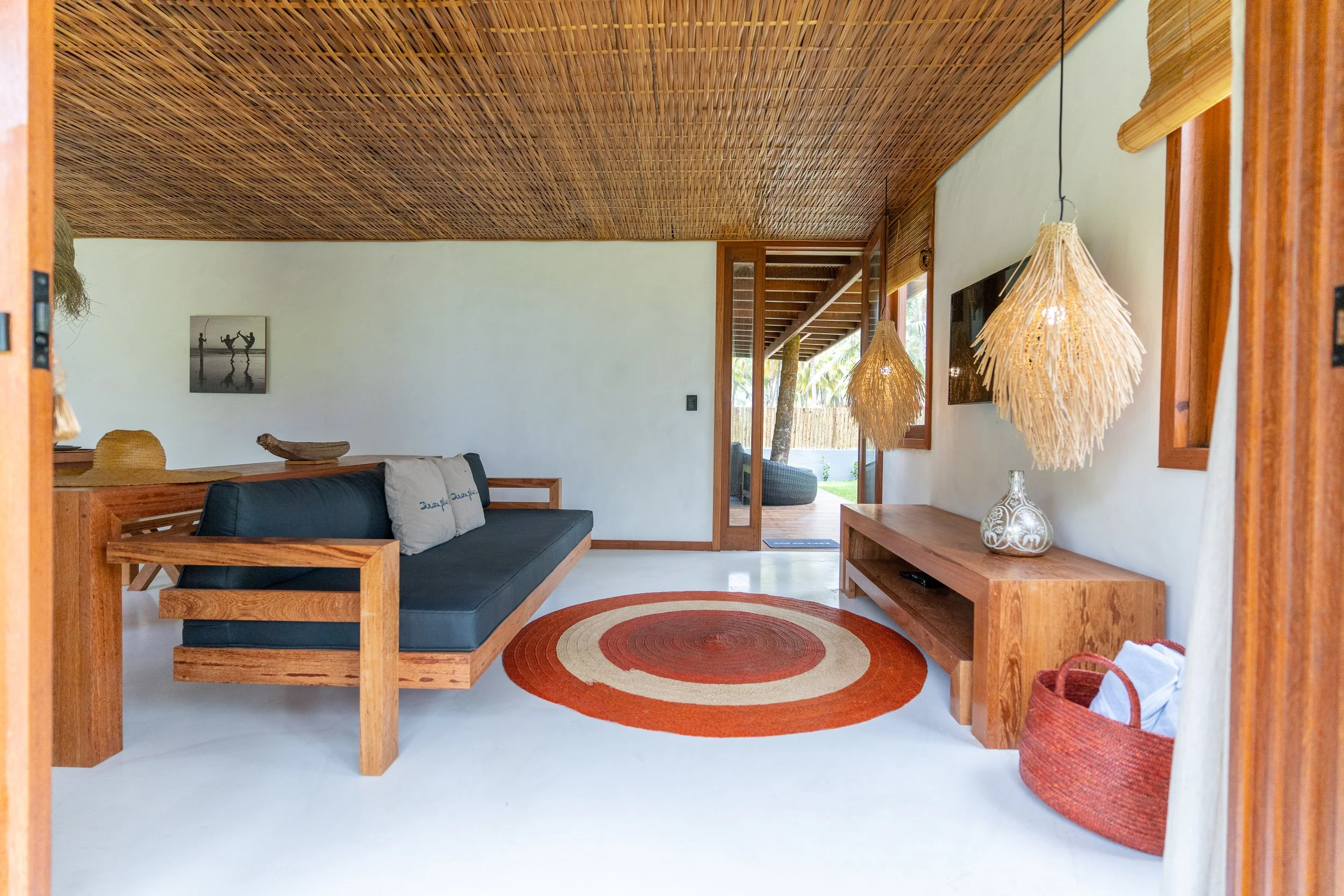 Sala de estar decorada de estilo natural com móveis de madeira, sofás com almofadas, tapete circular com listras verdes, lãs naturais penduradas do teto, parede branca com quadro de foto em preto e branco, porta de vidro que dá para varanda com vista