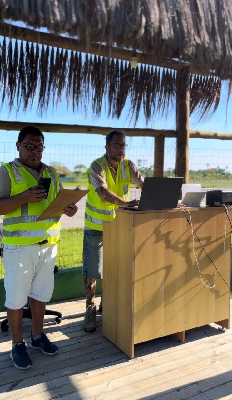 Homens trabalhando ao ar livre sob um quiosque com cobertura de palha, usando coletes de segurança amarelos. Um deles está usando um computador portátil na bancada de madeira, enquanto o outro verifica uma lista ou documento e usa o telefone móvel.