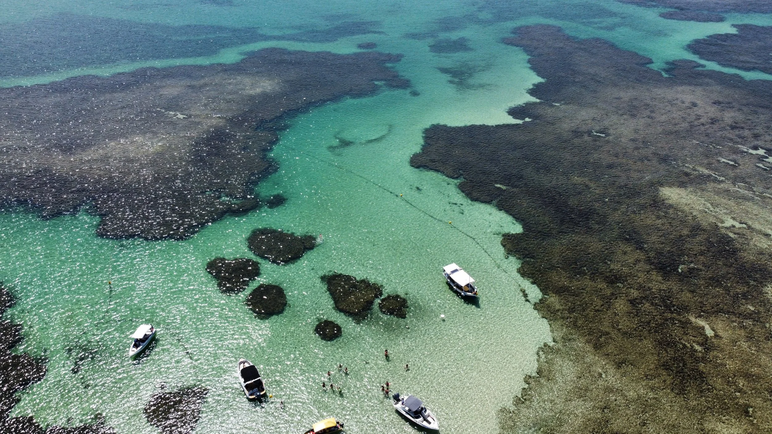 Imagem aérea de uma lagoa de água transparente com várias embarcações e pessoas nadando, rodeada por áreas de vegetação