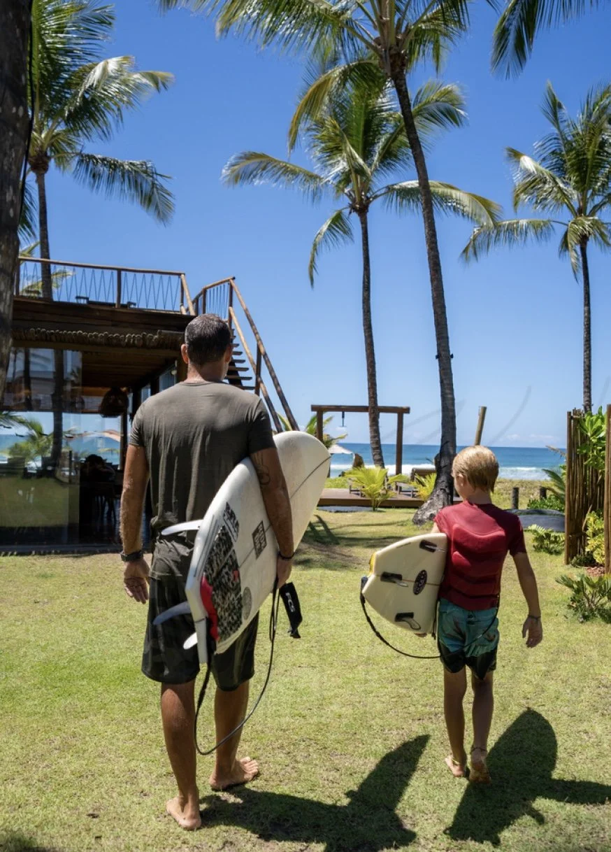 Pai e filho carregando pranchas de surfe em um jardim à beira-mar, com coqueiros e céu azul ao fundo.