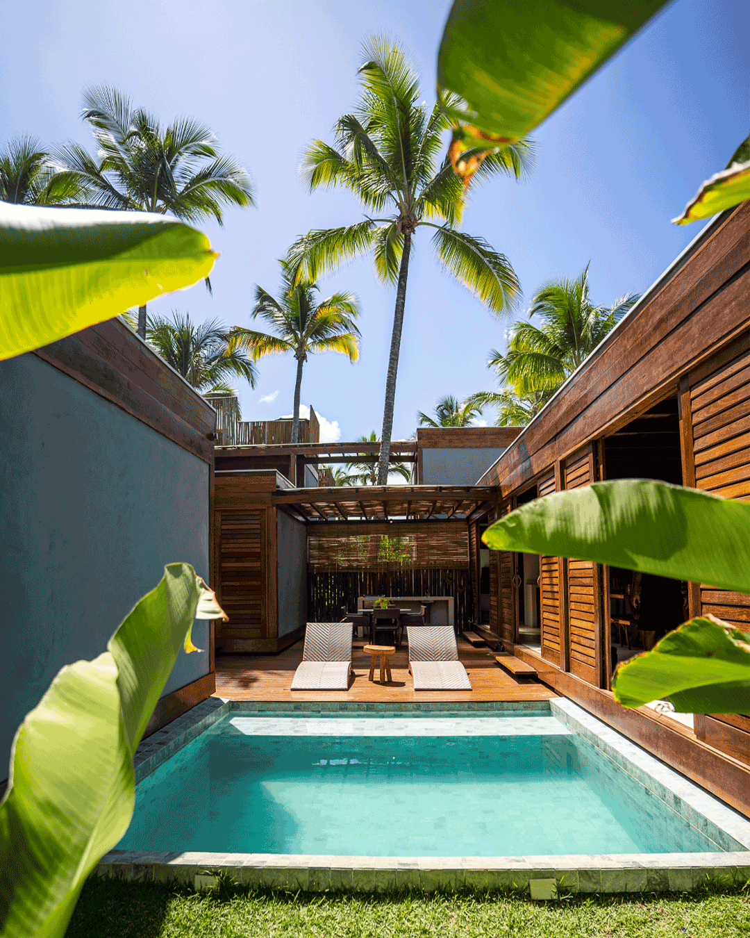 Piscina em um resort com espreguiçadeiras, área de lazer com móveis de madeira e palmeiras ao fundo.