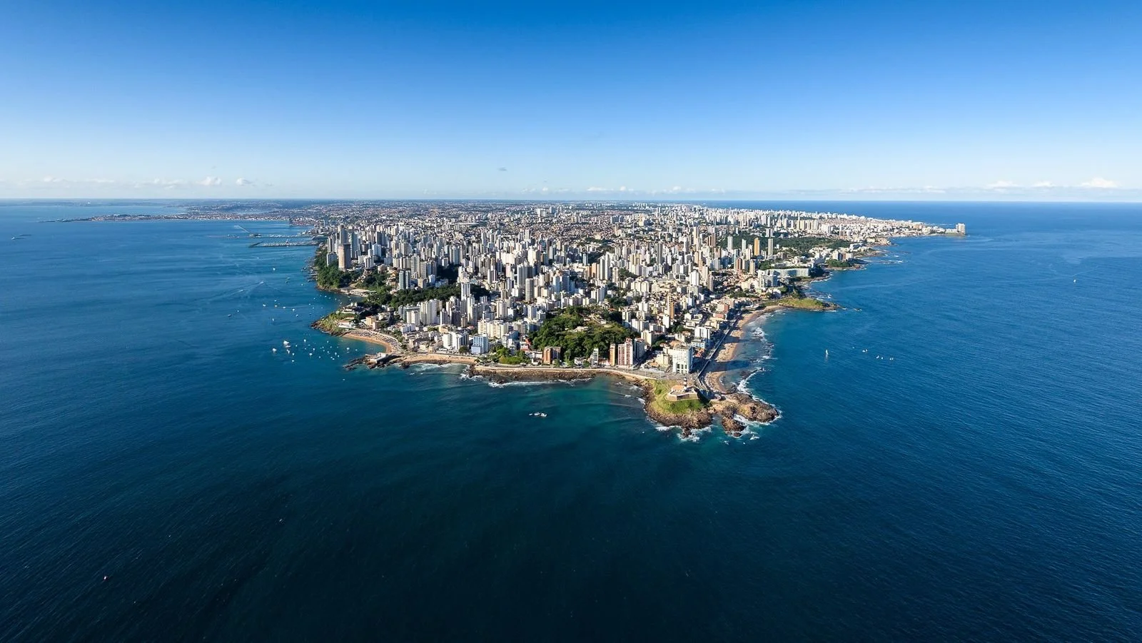 Vista aérea da orla de Salvador com muitos edifícios altos e praia, ao redor do oceano azul.