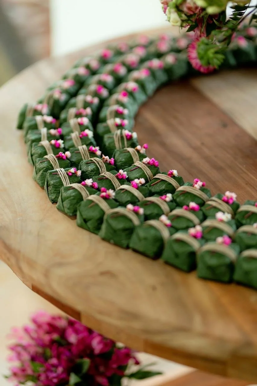 Trança de folha de bananeira com pequenos doces embrulhados em folha de bananeira, decorados com pequenos detalhes rosa e branco.