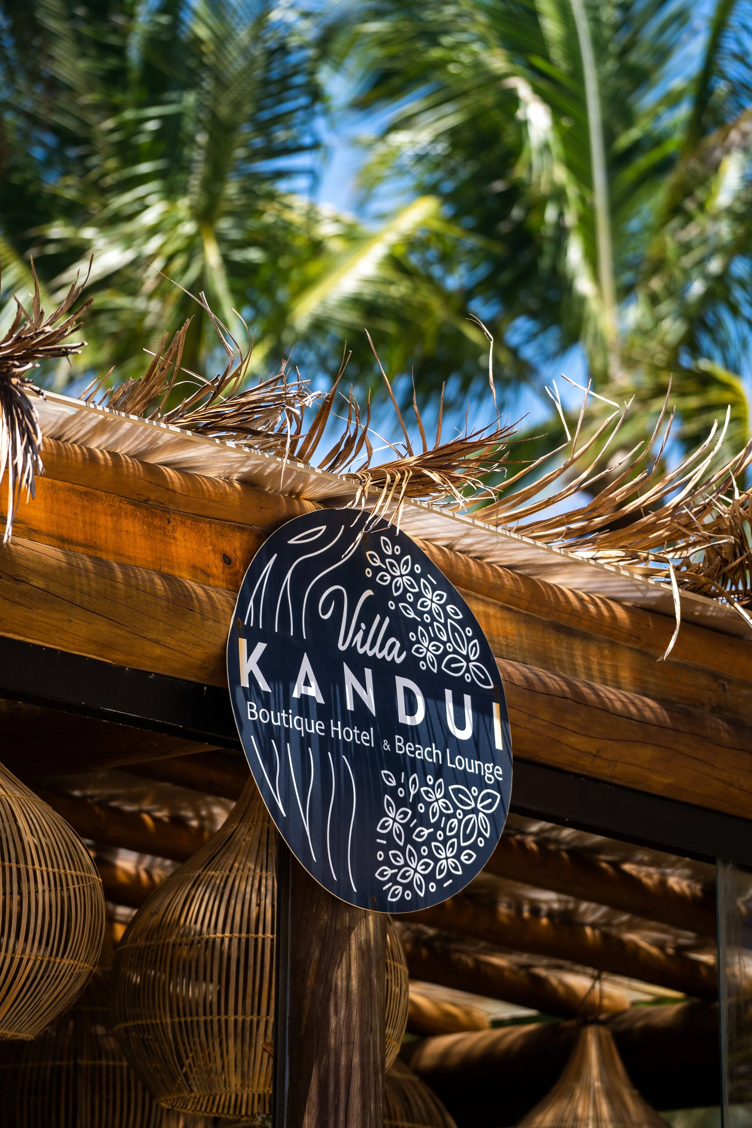 Placa do Villa Kandui Boutique Hotel & Beach Lounge em uma estrutura de madeira com folhas secas, rodeada por palmeiras ao fundo.