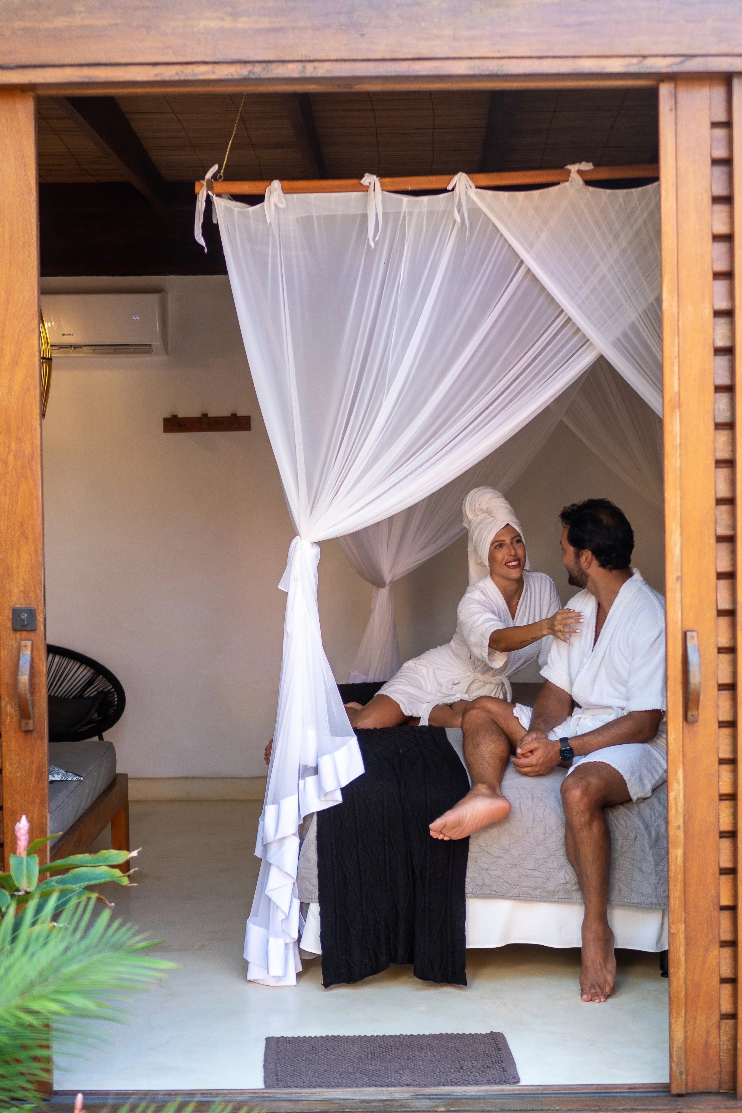 Casal sentado na cama com um mulher usando toalha na cabeça e um homem usando roupão branco, em quarto com cortina tipo cortina de mosquito, dentro de uma cabana de madeira.