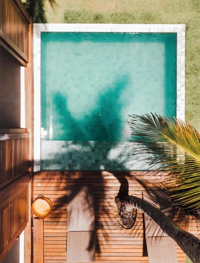 Vista aérea de uma piscina retangular ao lado de um deque de madeira com espreguiçadeiras e uma palmeira. A área ao redor é gramada.