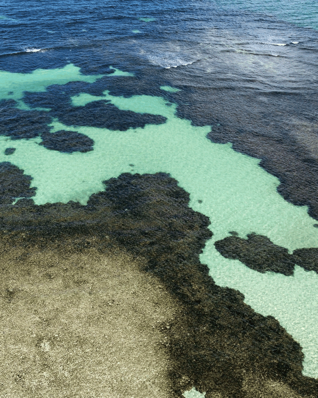 Água cristalina e marés de cores diferentes na costa, com evidências de formação de piscinas naturais e áreas de pedra