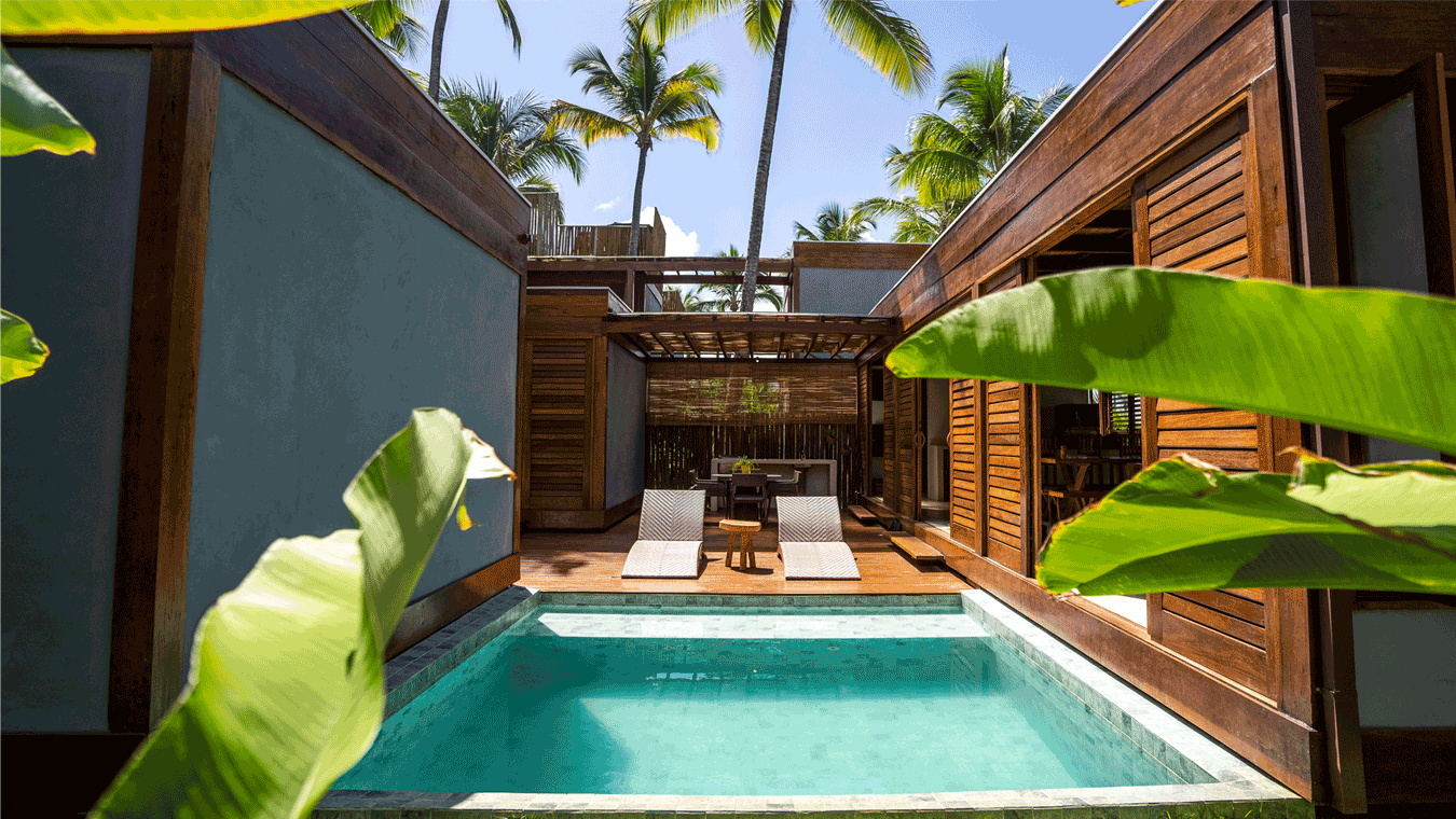 Pátio de uma casa de praia com piscina, cadeiras de descanso, mesas e cadeiras ao ar livre, cercado por paredes de madeira e plantas verdes, com coqueiros ao fundo e céu azul.