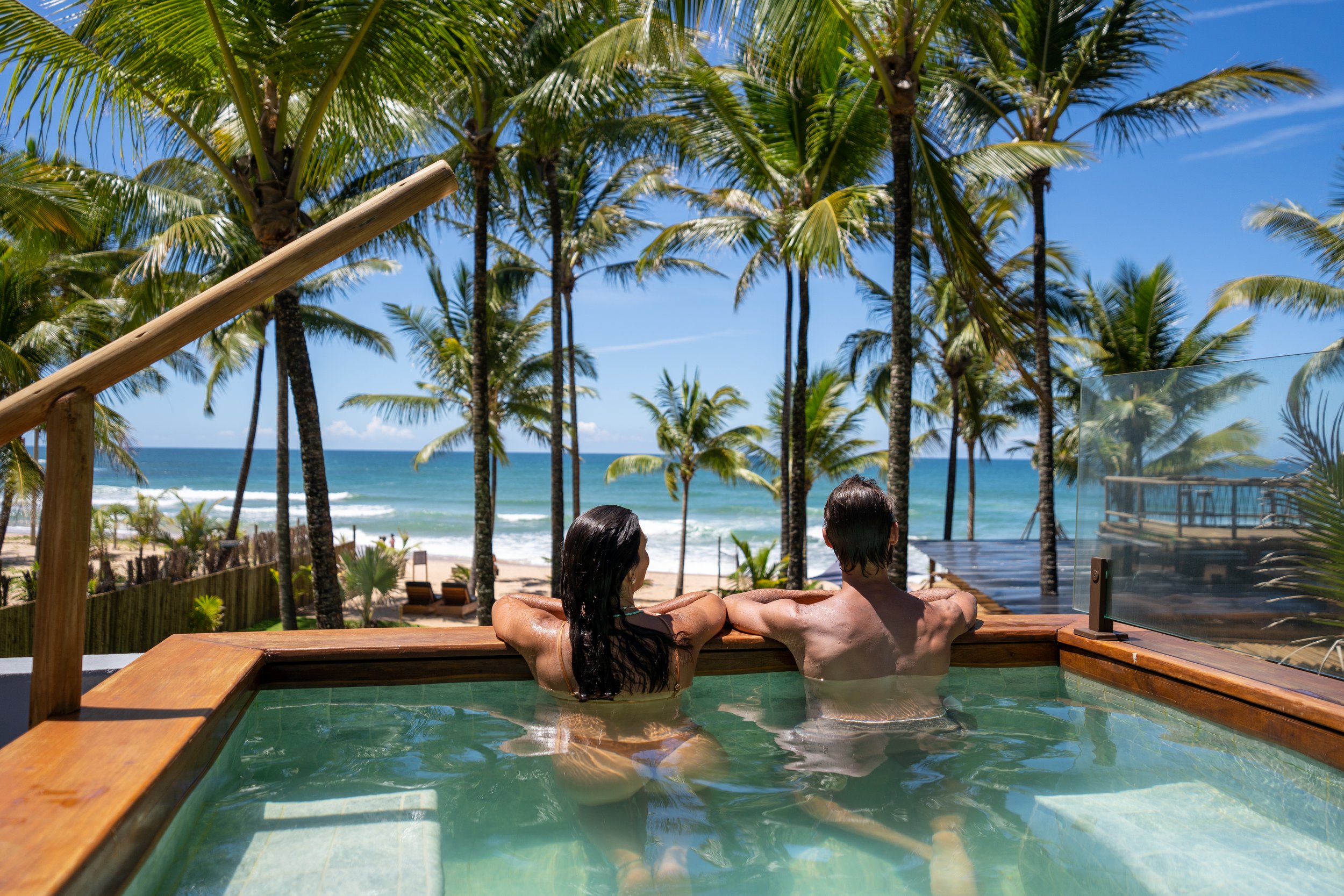 Casal relaxando em uma banheira de hidromassagem com vista para o mar e coqueiros sob céu azul.