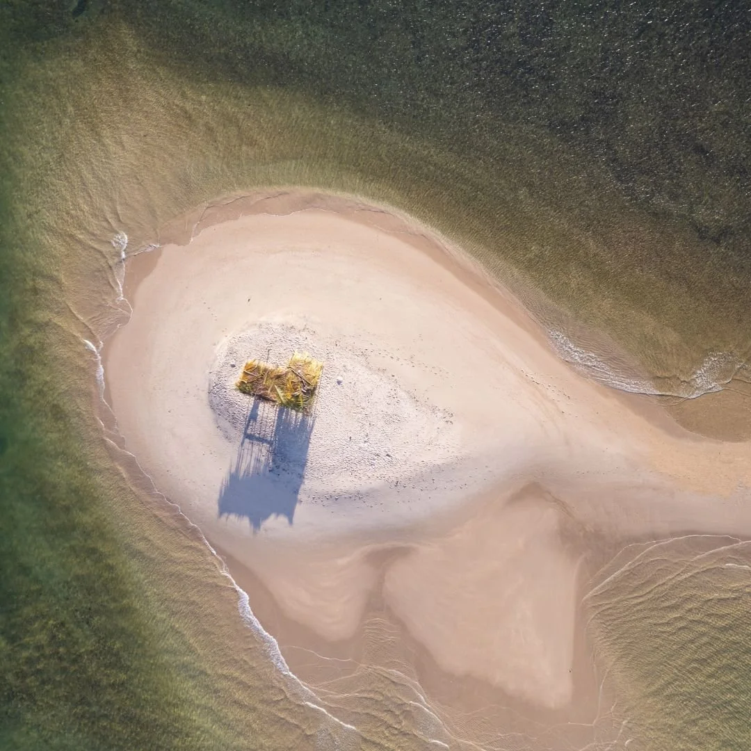 Vista aérea de uma pequena ilha de areia cercada por água, com uma estrutura ou cabana coberta de palha no centro. As ondas do mar tocam suavemente a praia ao redor da ilha.
