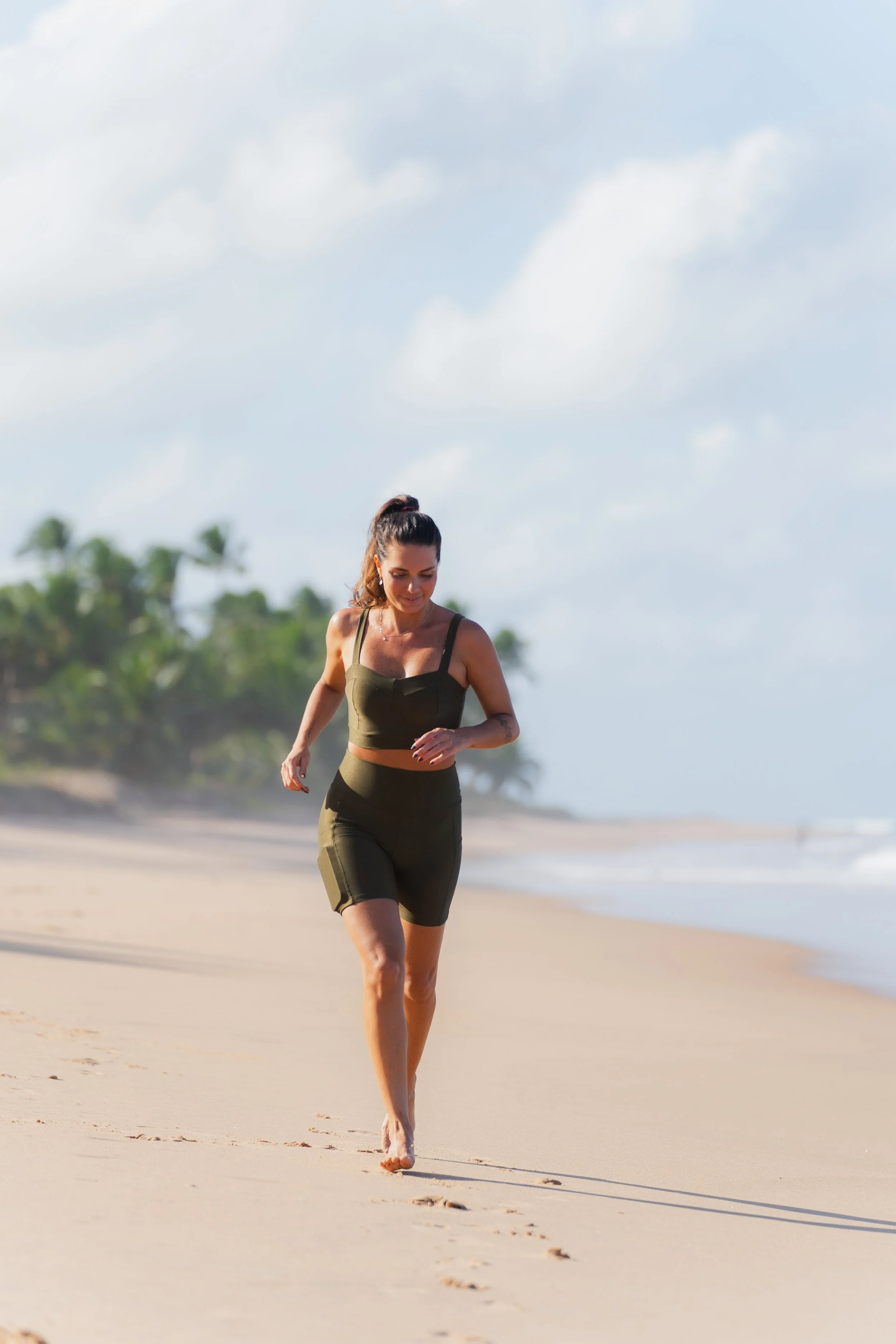 Mulher correndo descalça na praia, usando roupa esportiva verde, com coque de palmeiras ao fundo e céu parcialmente nublado.