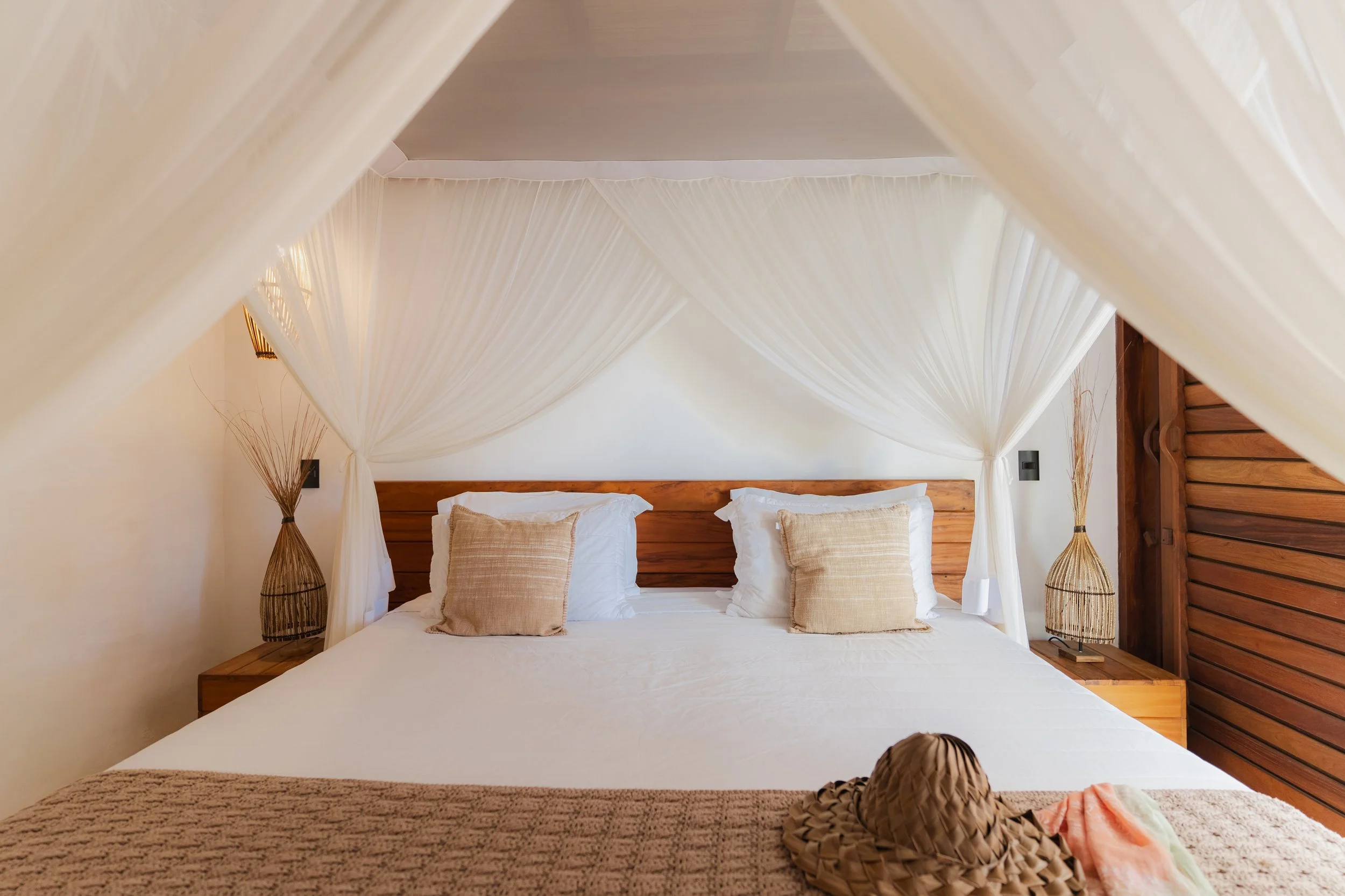 Cama de casal com dossel de cortina branca, almofadas bege, cabeceira de madeira, entre duas luminárias de mesa decorativas com vazados, paredes claras e detalhes de madeira.