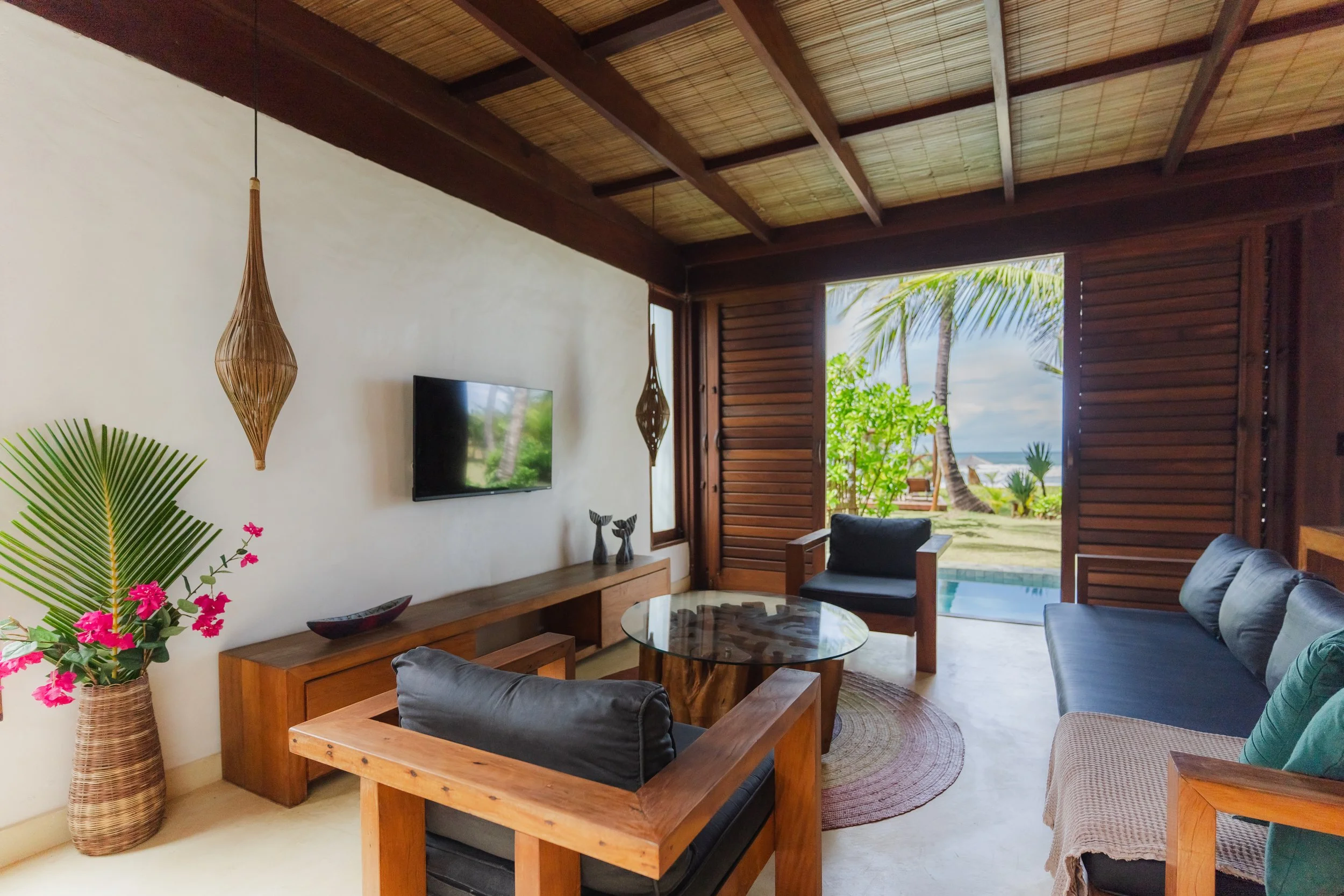 Sala de estar com móveis de madeira, poltronas de estilo tropical, paredes brancas, janelas de vidro, vista para a praia com coqueiros, plantas decorativas, iluminação natural.