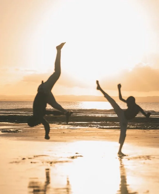Duas pessoas praticando capoeira em uma praia ao pôr do sol, com movimentos acrobáticos.