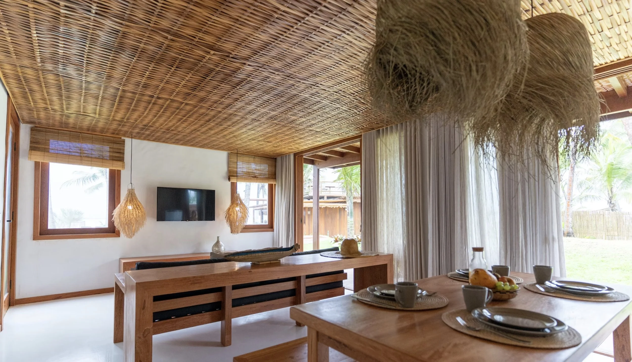 Sala de jantar com móveis de madeira, paredes claras, janelas com persianas de bambu, iluminação com luminárias de fibra natural, decoração tropical com vasos, para-sóis e plantas visíveis através das janelas.