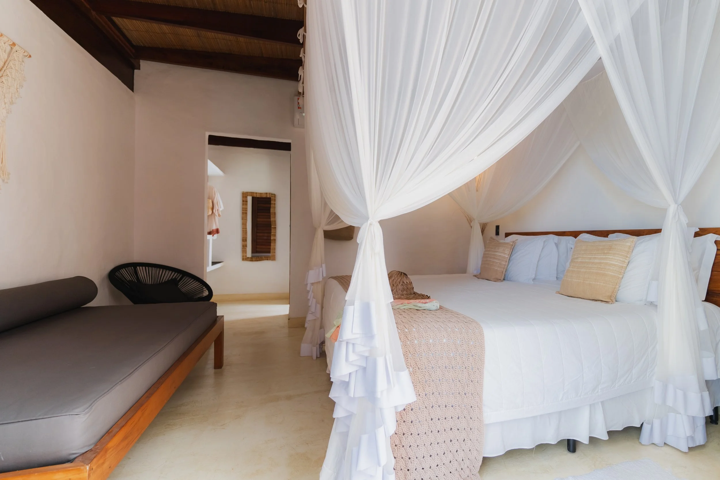 Quarto com cama de casal com cortina de dossel branca, travesseiros bege, parede branca, painel de madeira, espelho retangular na parede, sofá cinza e cadeira preta de fibras.
