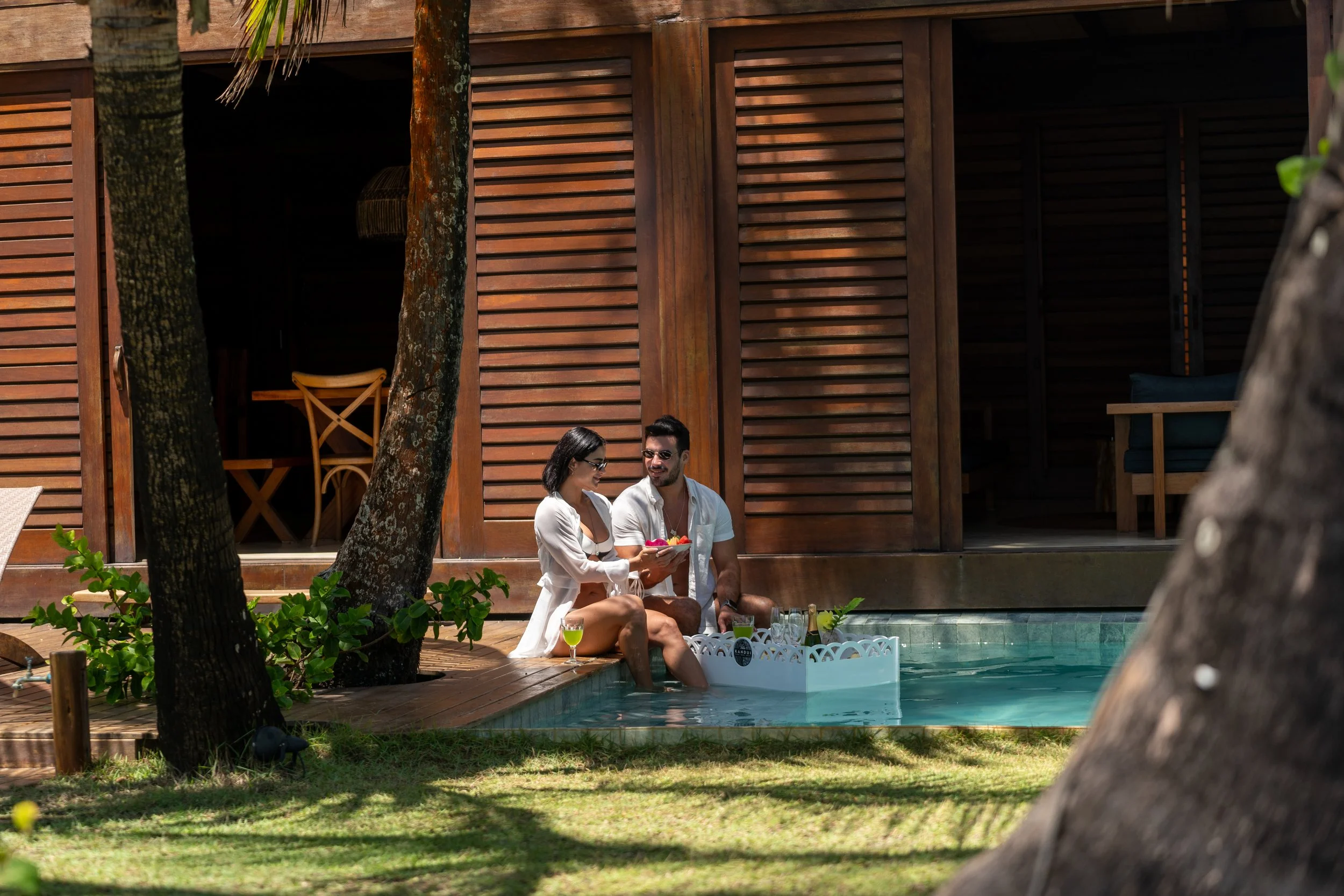 Casal sentado na beira de uma piscina, com o homem sorrindo e a mulher segurando uma tigela de comida, ao lado de uma cabana de madeira, rodeados por árvores.
