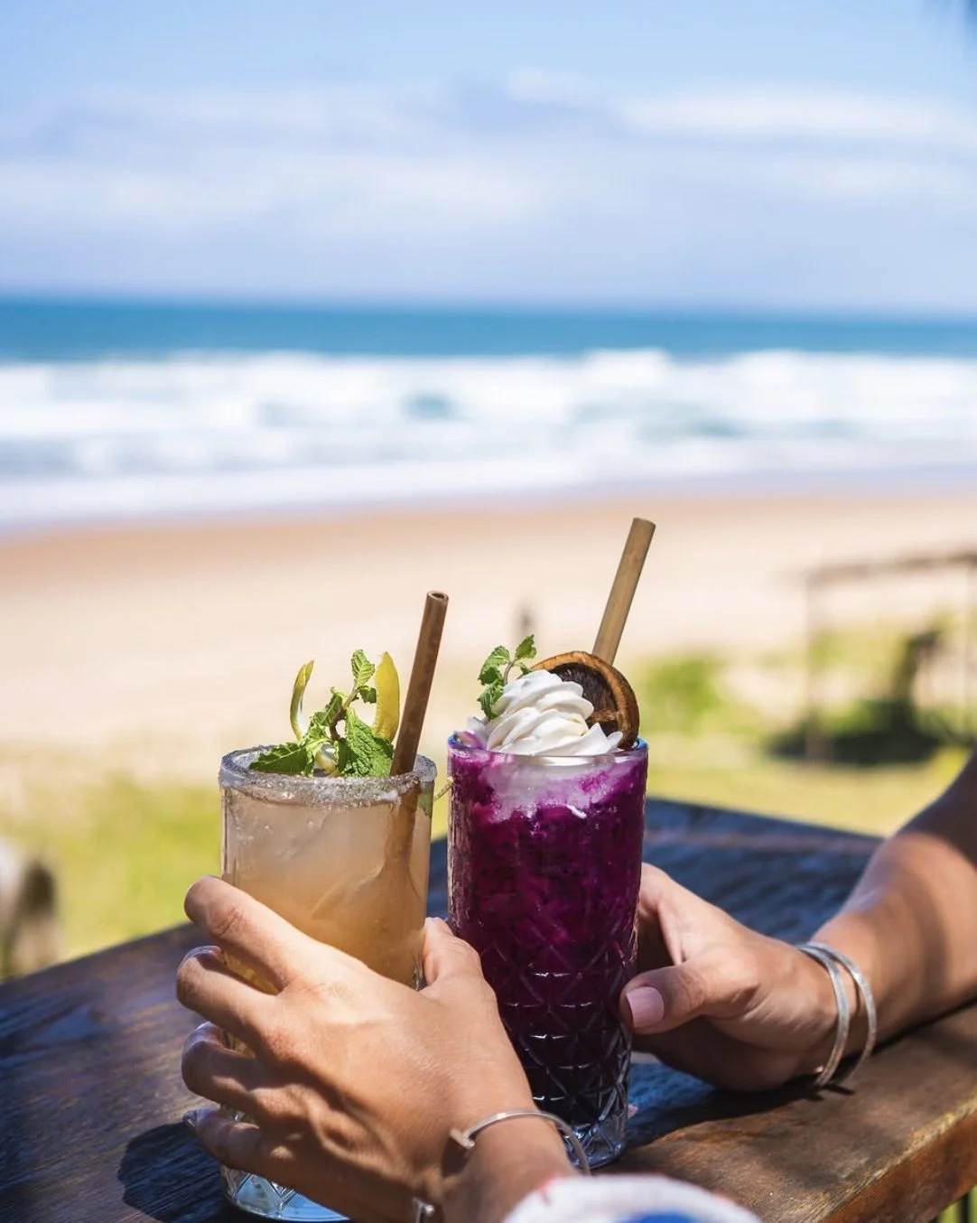 Duas pessoas brindam com drinks coloridos à beira da praia, com o mar ao fundo. Um drink é marrom claro com hortelã, e o outro é roxo com chantilly e fatia de fruta.