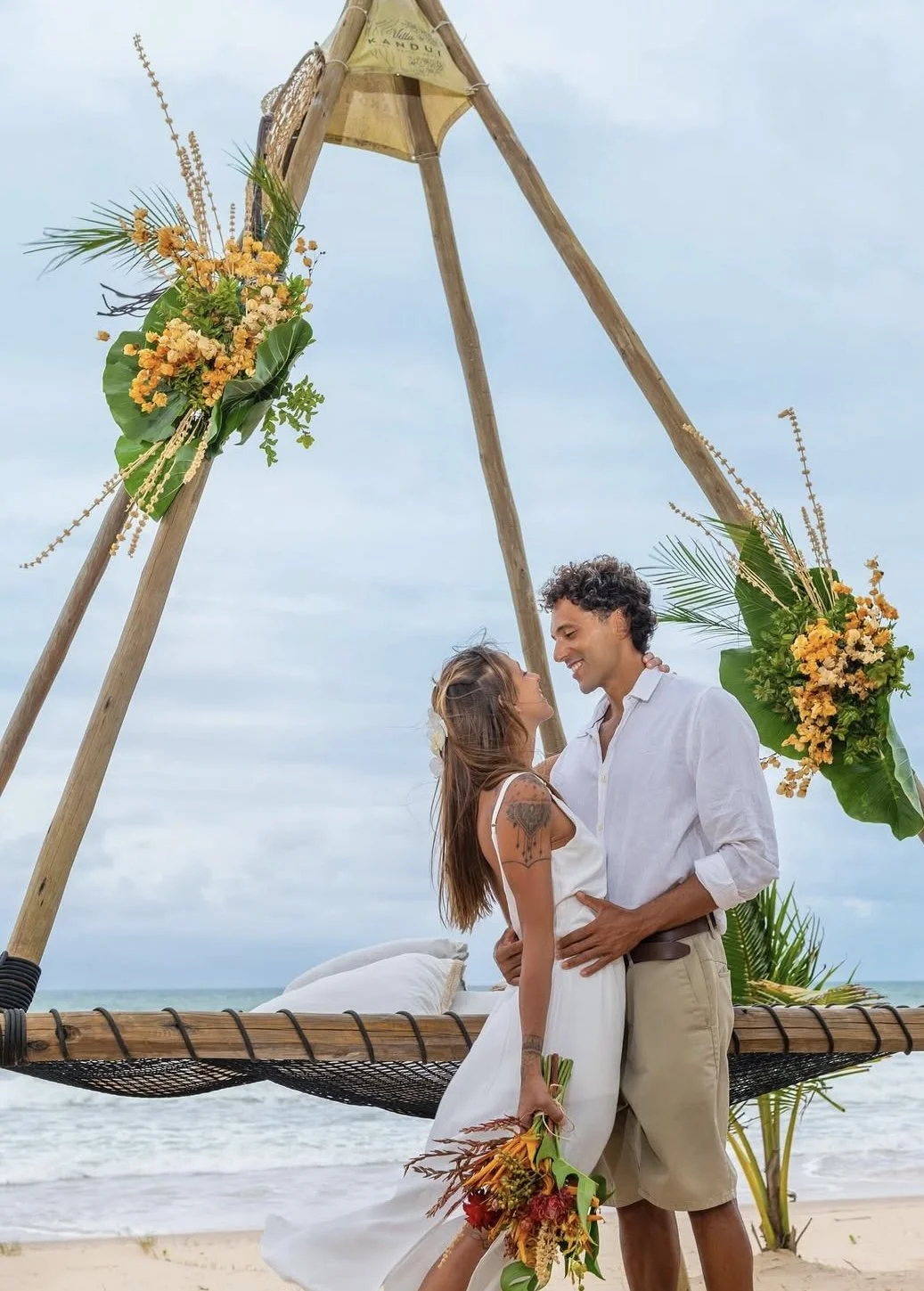 Casal se apaixonando na praia durante casamento ao ar livre, com decoração de flores e uma estrutura de madeira.