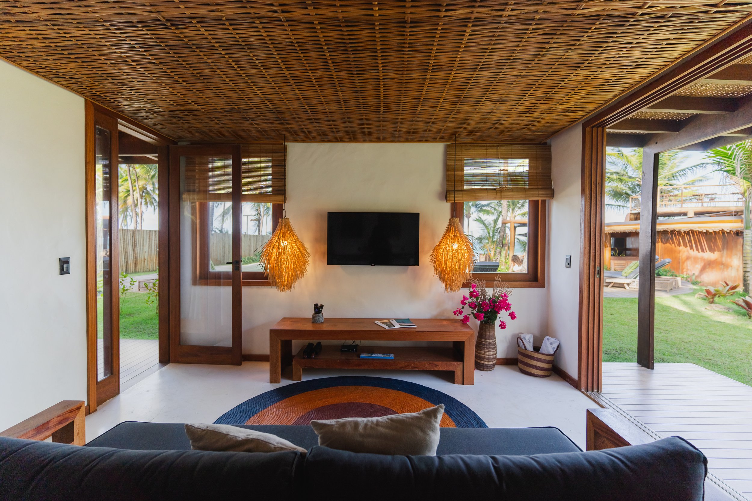 Sala de estar com móveis de madeira, janela grande com vista para jardim, luminárias pendentes de sisal, plantas e decoração aconchegante